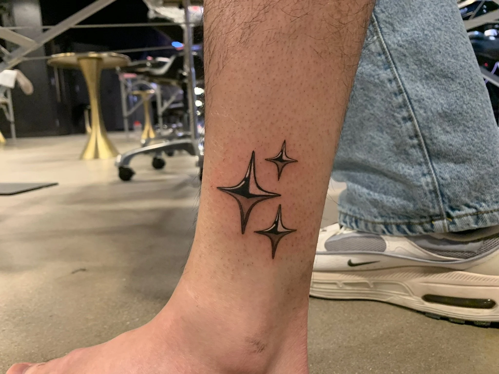 Tattoo of four black outlined star shapes on person's lower leg, with the person kneeling on one knee, wearing light blue denim shorts and gray Nike sneakers, in what appears to be an indoor gym or workout area.