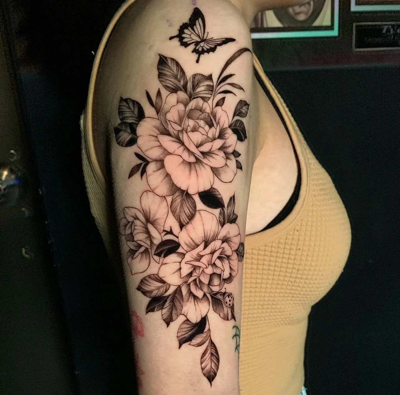 Black and gray floral tattoo featuring large roses, leaves, a butterfly, and a ladybug on the upper arm of a person wearing a yellow sleeveless top.