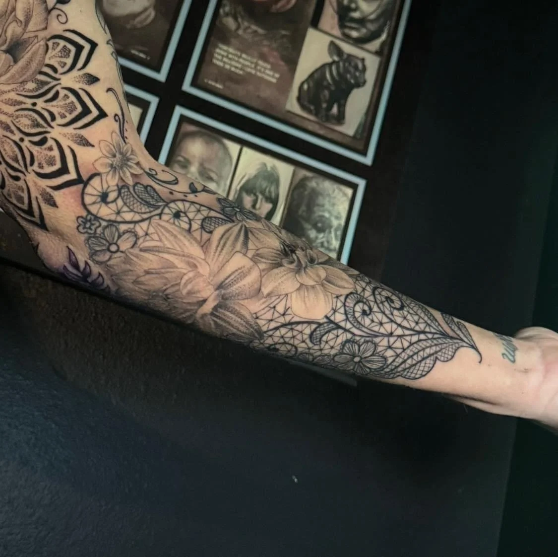 Tattooed arm with floral and geometric patterns, and a small part of a tattoo visible near the wrist, with framed portraits and artwork on the wall in the background.
