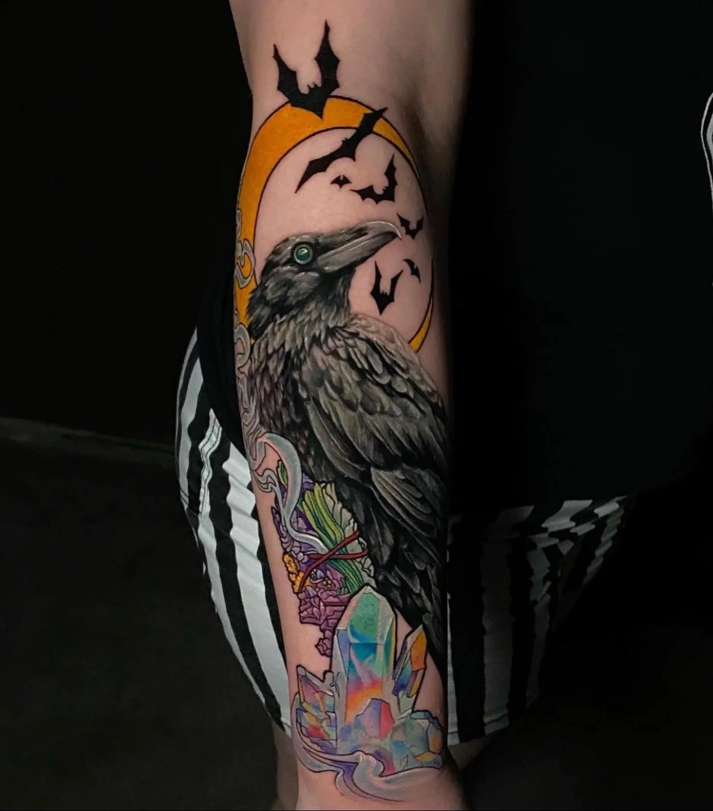 Tattoo of a raven with detailed feathers, a green eye, surrounded by bats, a moon, colorful crystalline formations, and a nautical rope, on person's leg.