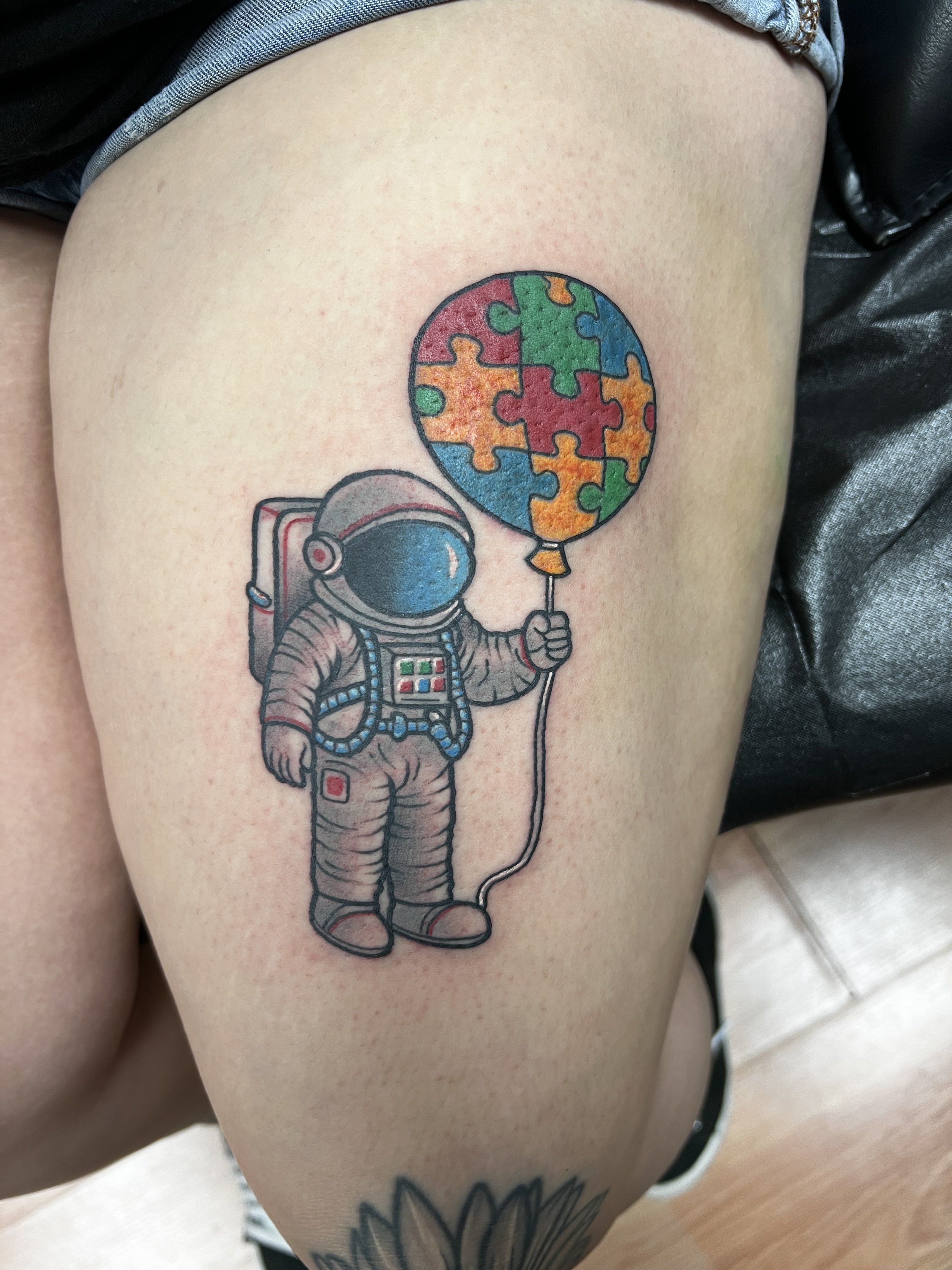 Tattoo of an astronaut holding a balloon made of puzzle pieces on a person's thigh.