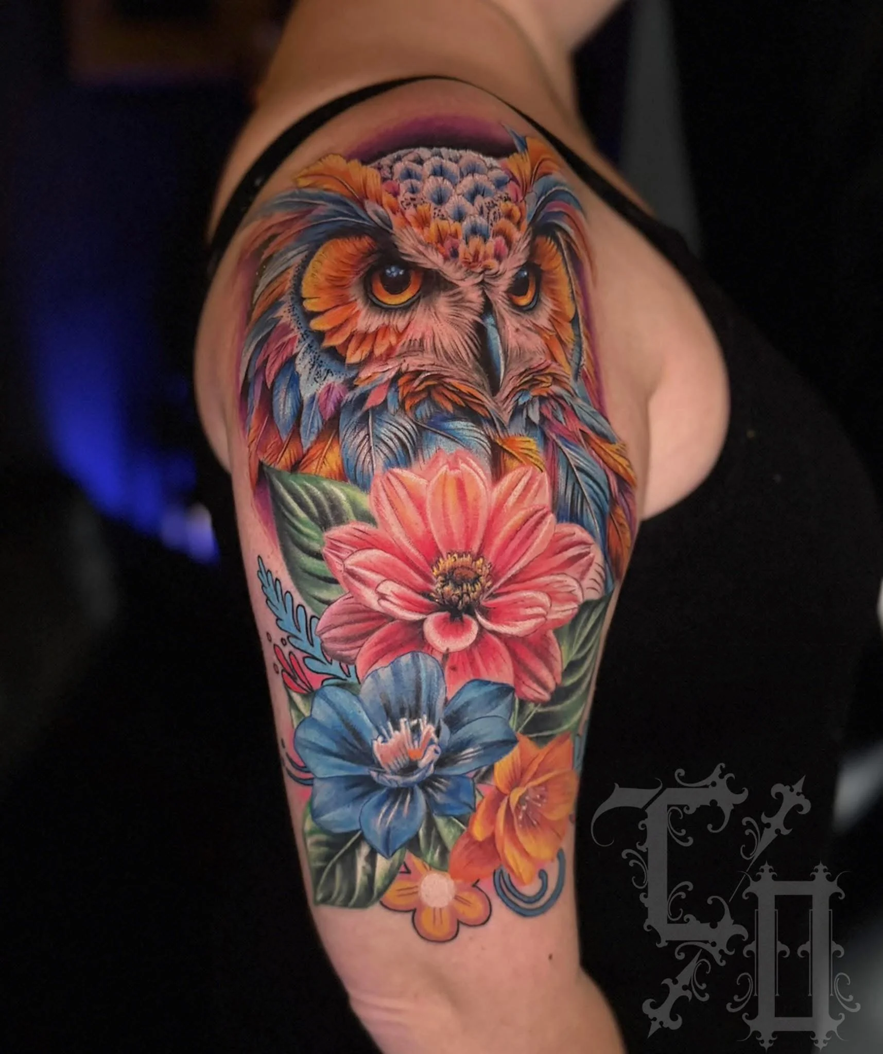 Colorful tattoo of an owl with detailed feathers above vibrant pink and blue flowers on a person's upper arm.