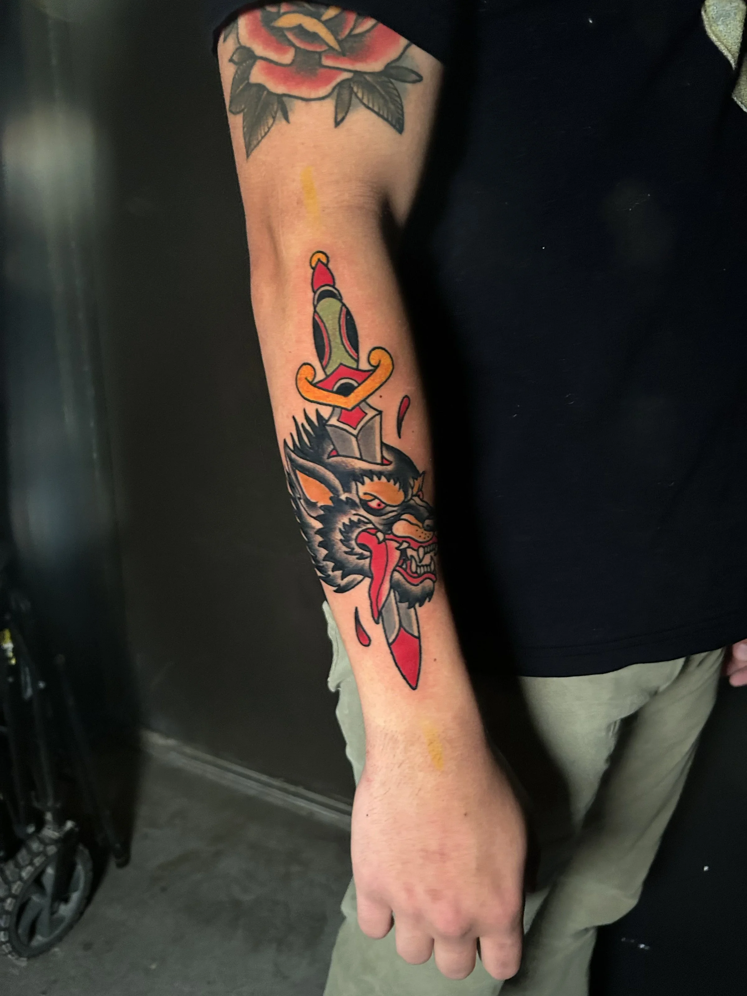 a traditional style tattoo of a black wolf with a sword going through the top of the wolf's head in black, yellow, red and green on a person's forearm