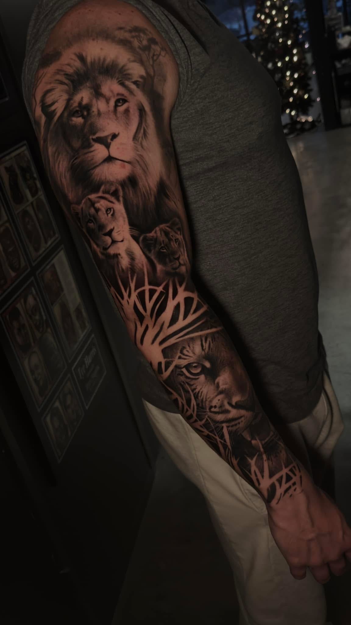 Close-up of a person's arm with a detailed black and gray tattoo sleeve featuring a lion's face at the top, a pair of lion cubs in the middle, and a lion's face near the wrist, with a graphic design resembling tree branches below