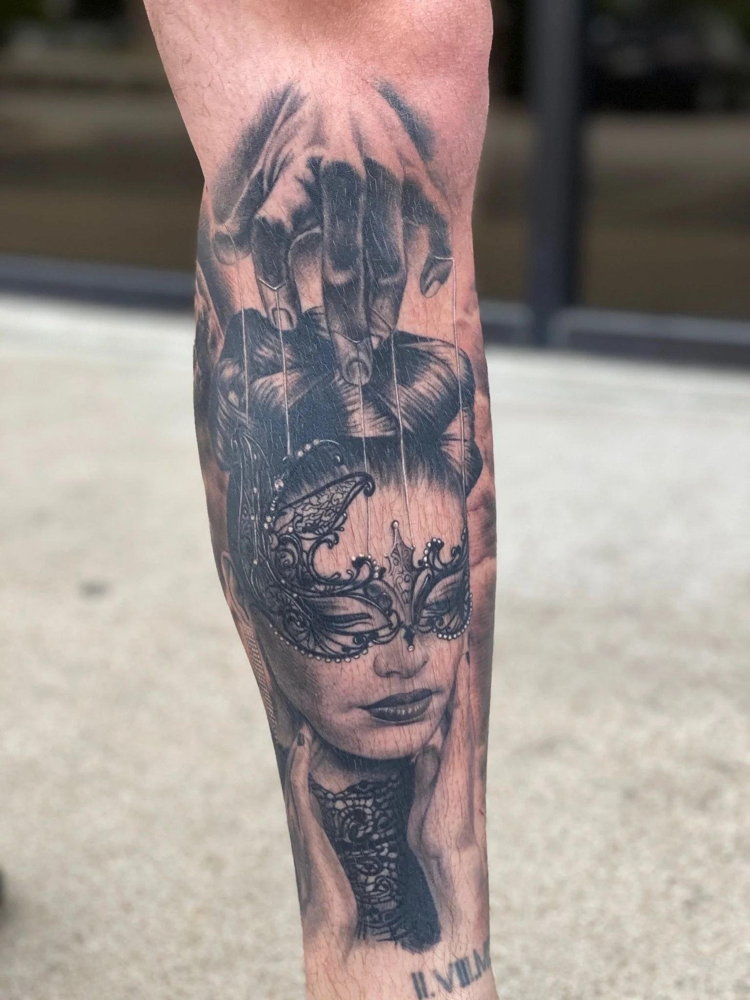 Black and gray tattoo of a woman's face with a butterfly mask covering her eyes, her hand on her cheek, and a hand above her head, with an intricate design and script below.