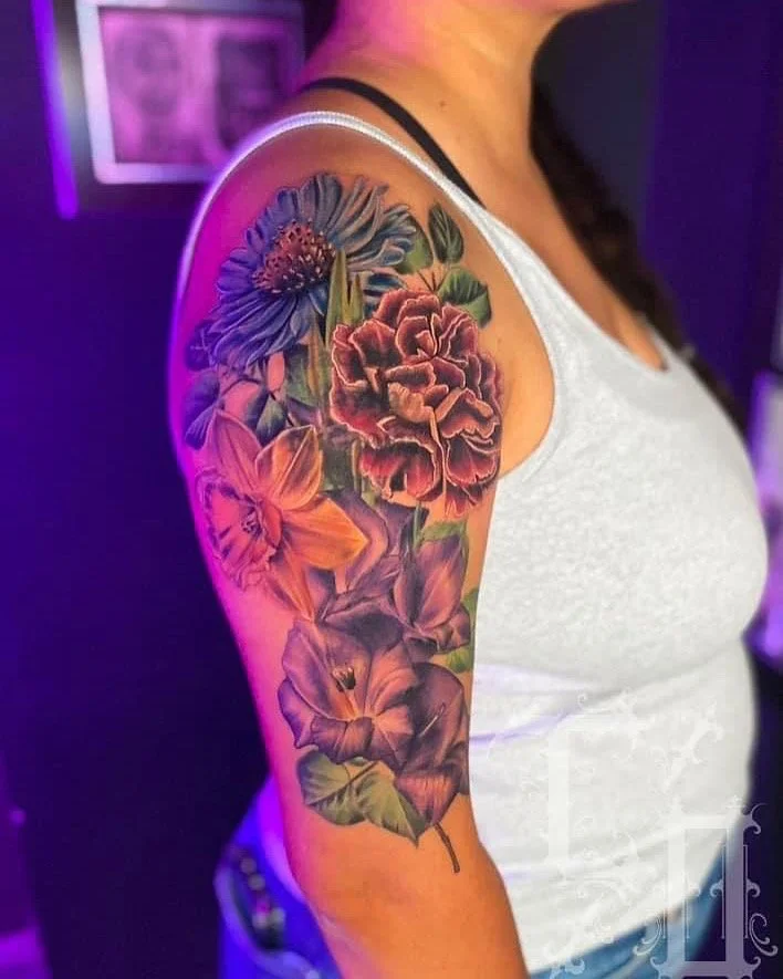 Colorful floral tattoo on woman's upper arm, featuring various flowers including a large red peony, purple and orange blossoms, and green leaves.