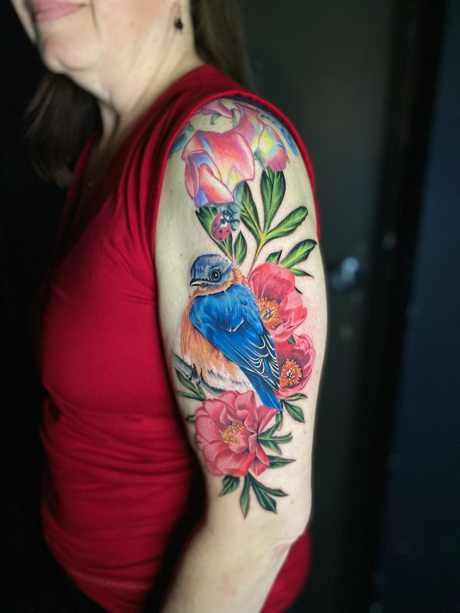 Colorful tattoo of a blue bird on a branch surrounded by pink and orange flowers and green leaves on a woman's upper arm.