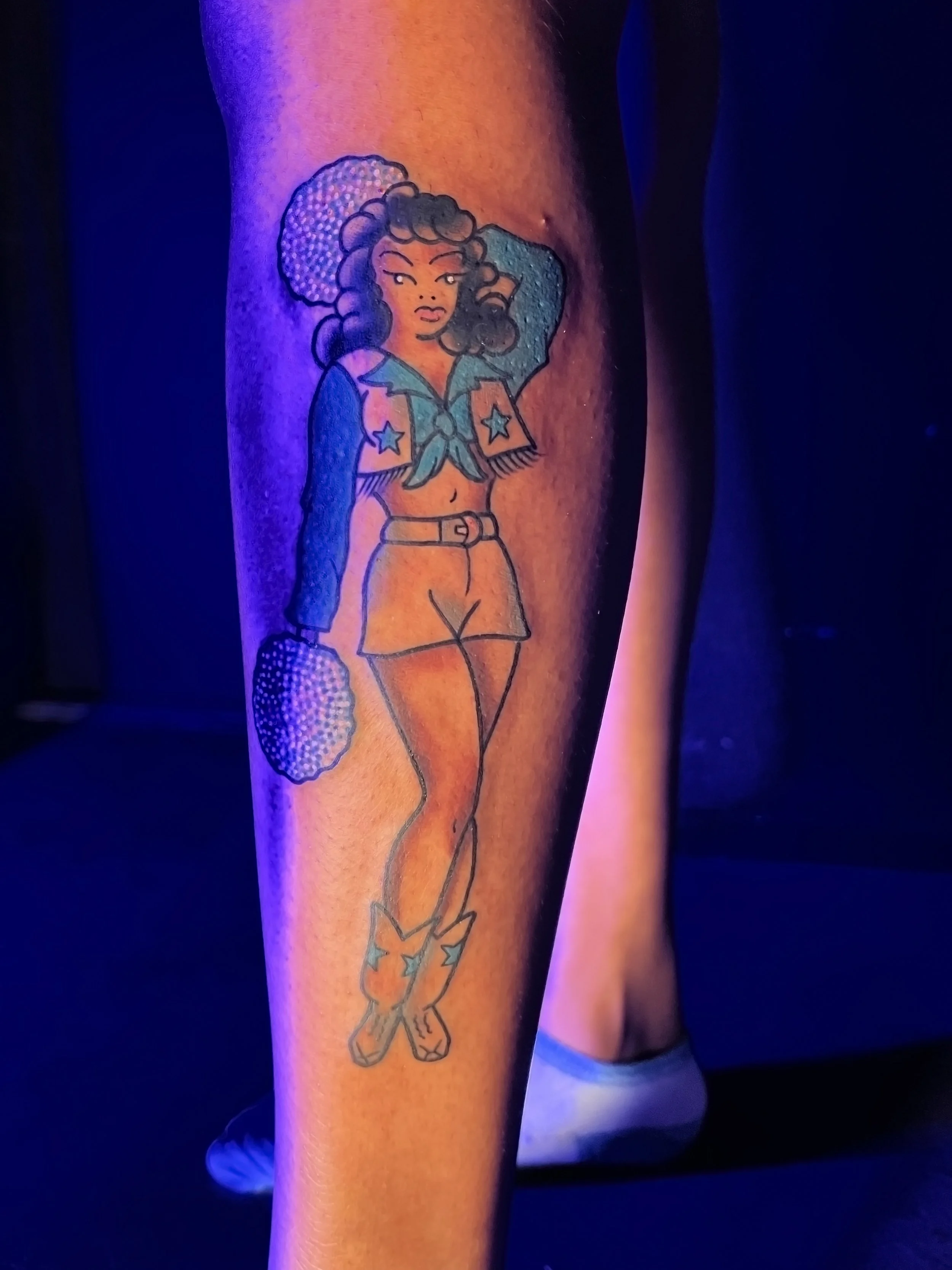 a colorful, traditional style black dallas cowboys cheerleader tattoo on a person's outside calf