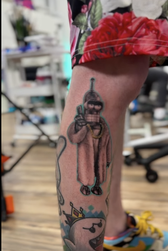 Tattoo of a character with a retro movie camera head, wearing a pink robe, holding a camera, on person's calf. Blender of colorful designs, possibly cartoonish, below.