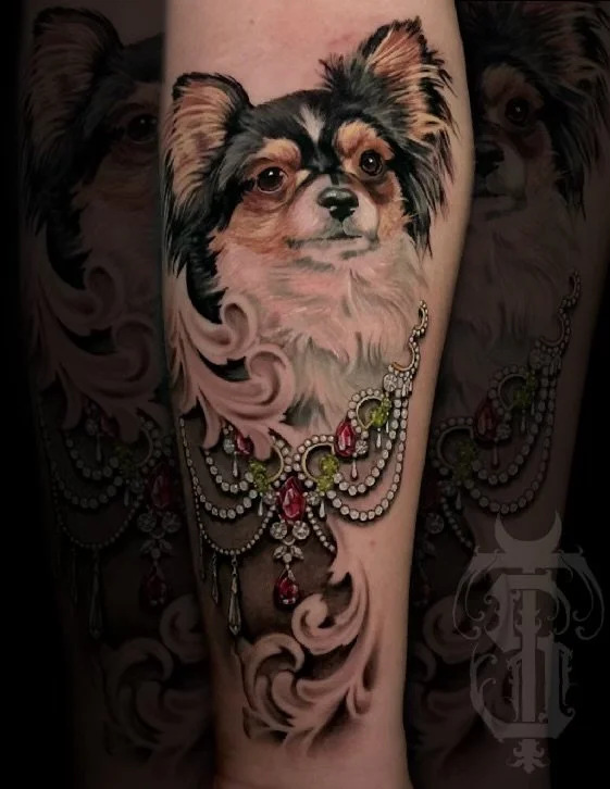 Tattoo of a small, fluffy dog with long ears, adorned with a pearl and gemstone necklace, on a person's arm.