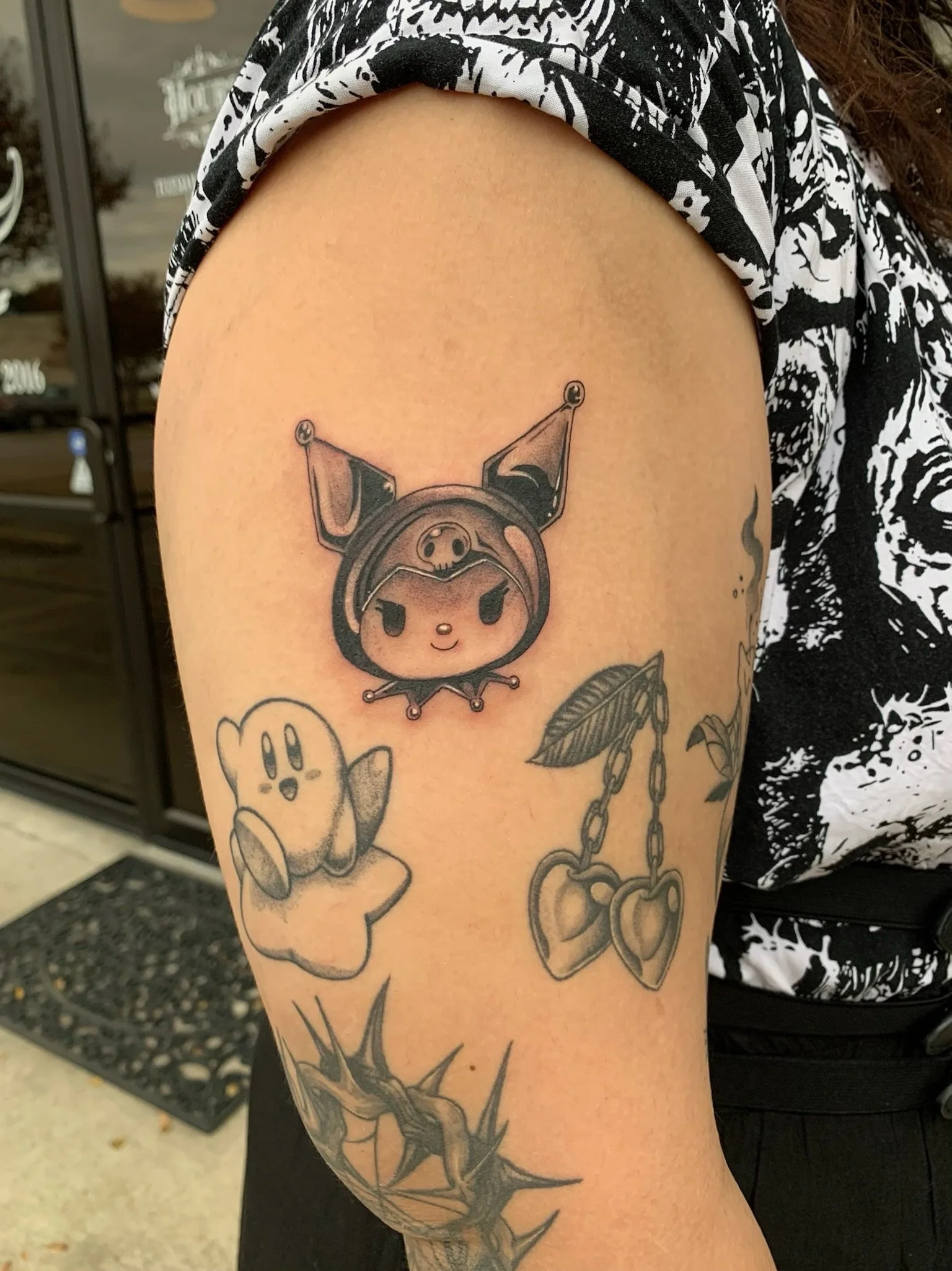 Tattoos on upper arm featuring characters and objects, including a cute fox or puppy with large ears, a ghost-like figure, and a pair of cherries with leaves.
