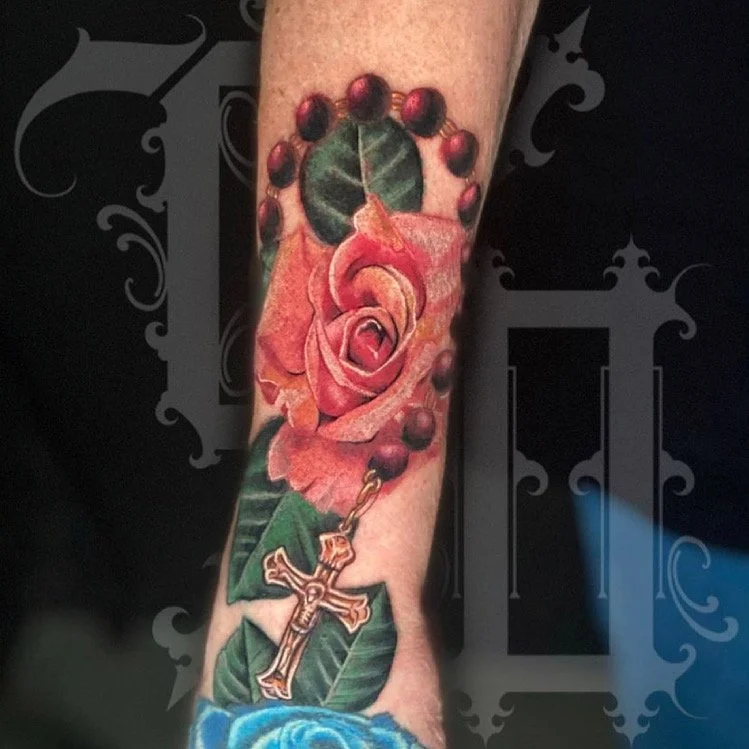 Tattoo of a pink rose with green leaves and a rosary with dark beads, a gold cross, and a chain hanging from the rose, on a person's arm.