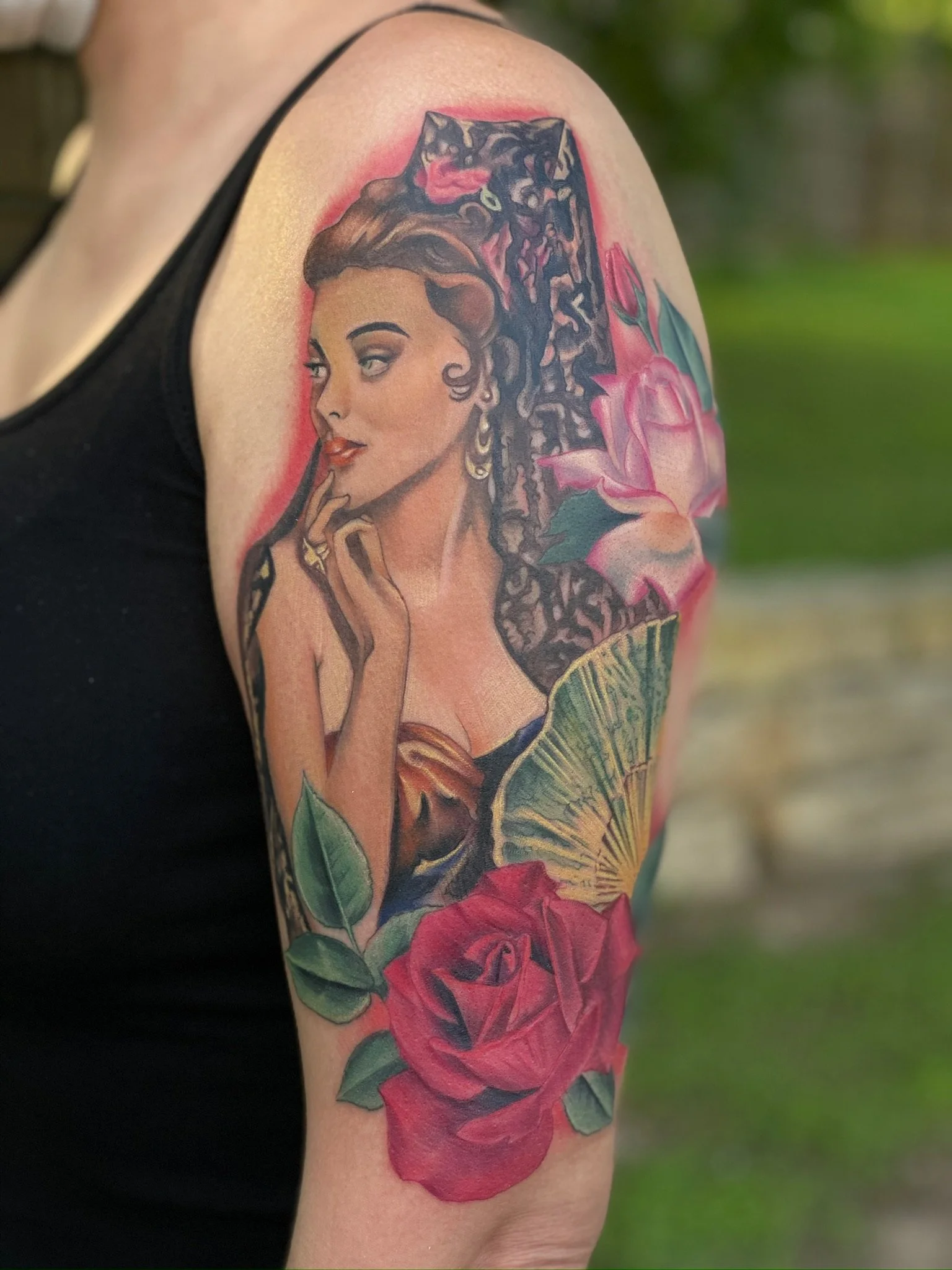 Tattoo of a vintage-style woman with a flower and leaves, partially covered by a pink rose and a green fan, on the upper arm.