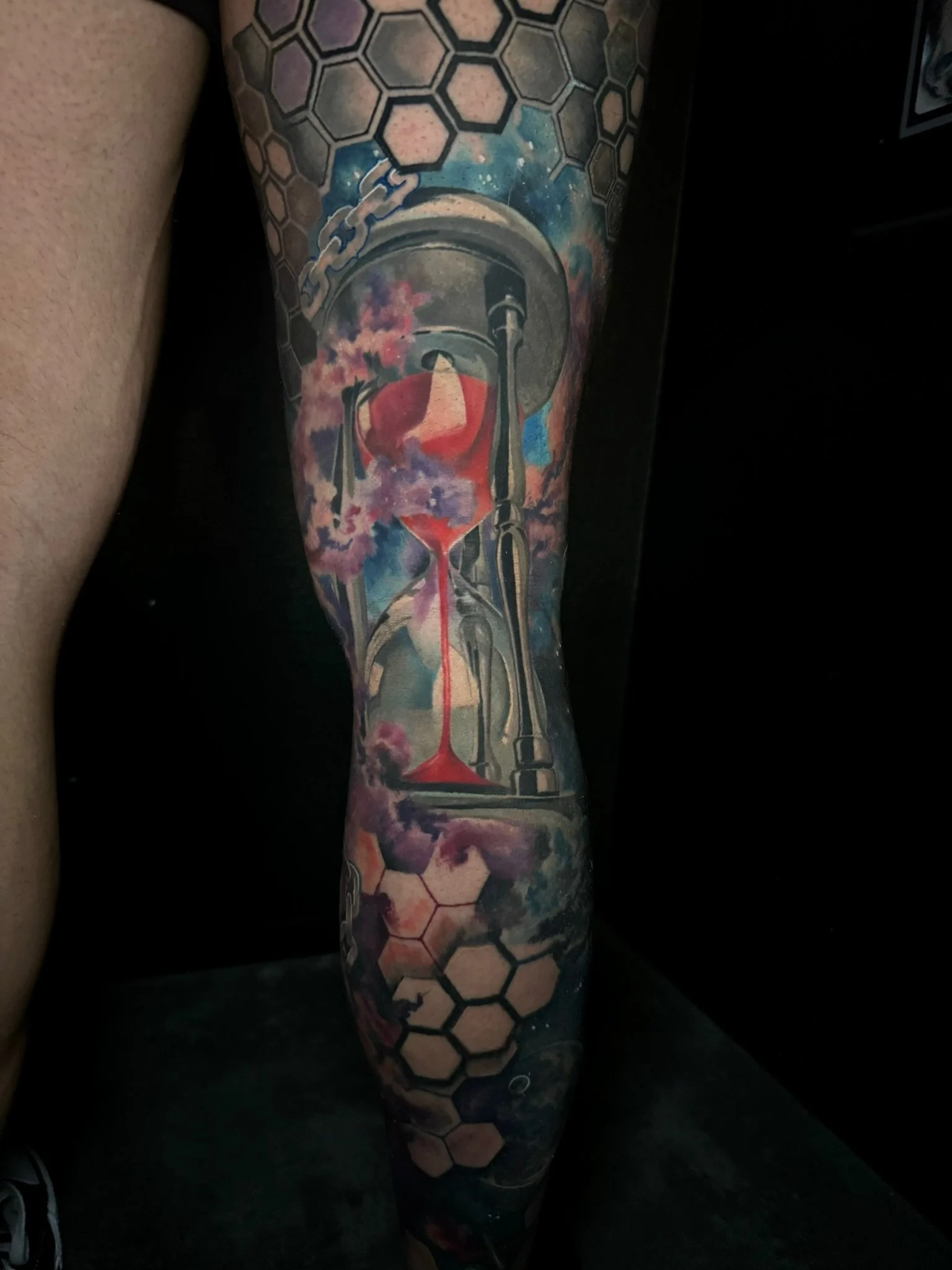 Colorful tattoo featuring an hourglass with pink and purple clouds, honeycomb patterns, and abstract cosmic elements.