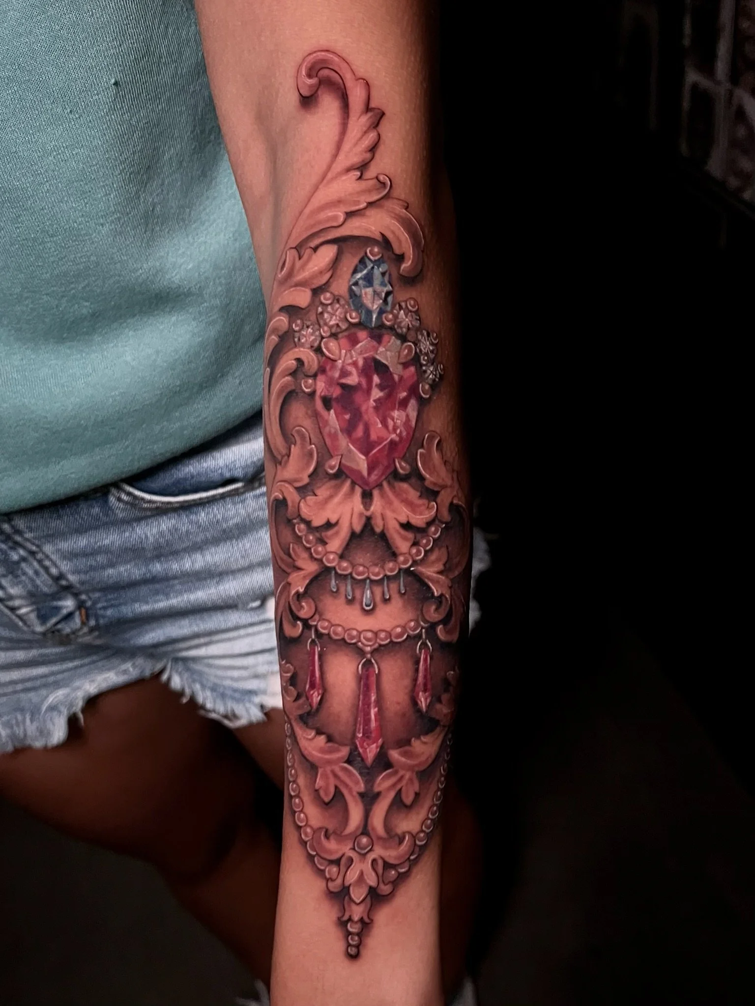 Colorful tattoo of a large red gemstone surrounded by pink and black decorative elements on a person's forearm.