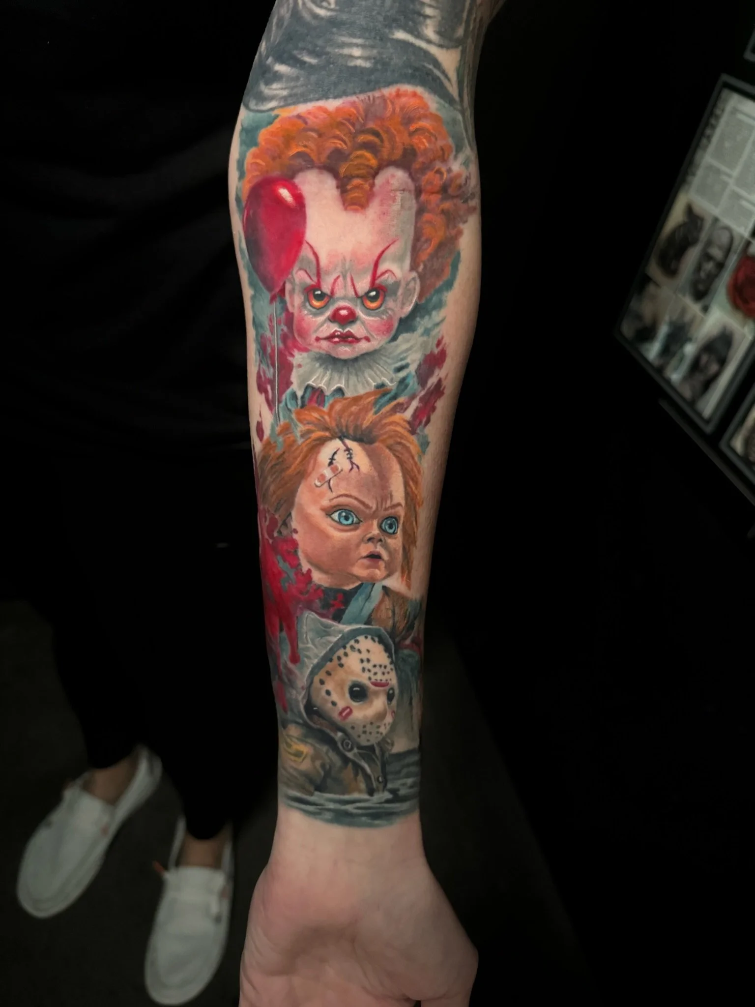 Colorful tattoo of horror movie characters on a person's forearm, featuring Pennywise the clown, Chucky doll, and Jason Voorhees from Friday the 13th.
