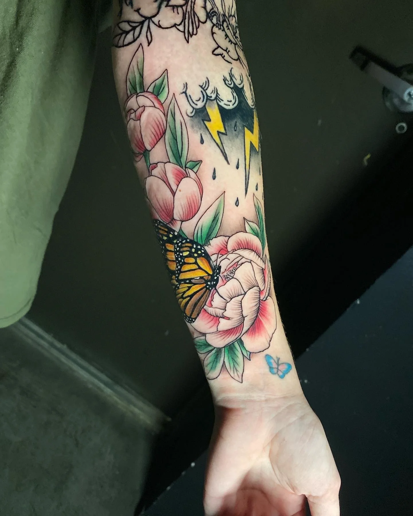 Colorful tattoo featuring pink peonies, a Monarch butterfly, a blue butterfly, dark clouds with lightning bolts, and raindrops.