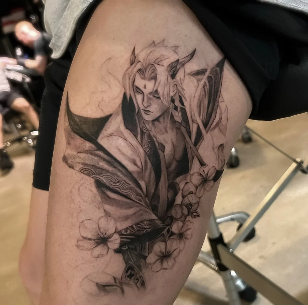 A detailed black and gray tattoo of an anime-style character with long hair, pointed ears, and a headband. The character is surrounded by flowers and has intricate patterns on their clothing.