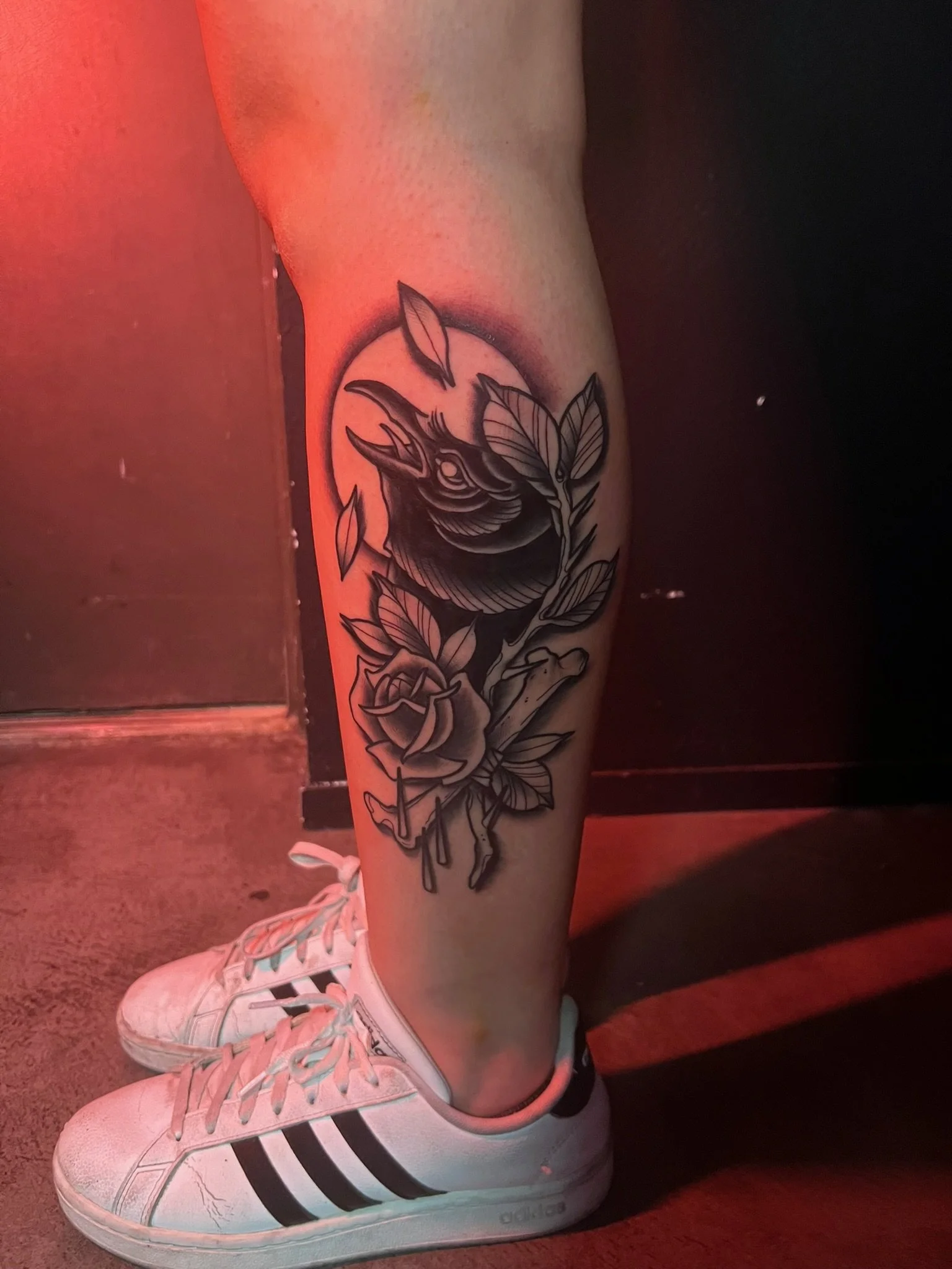 Tattoo of a crow surrounded by leaves and a rose on a person's lower leg, wearing white Adidas sneakers with black stripes.