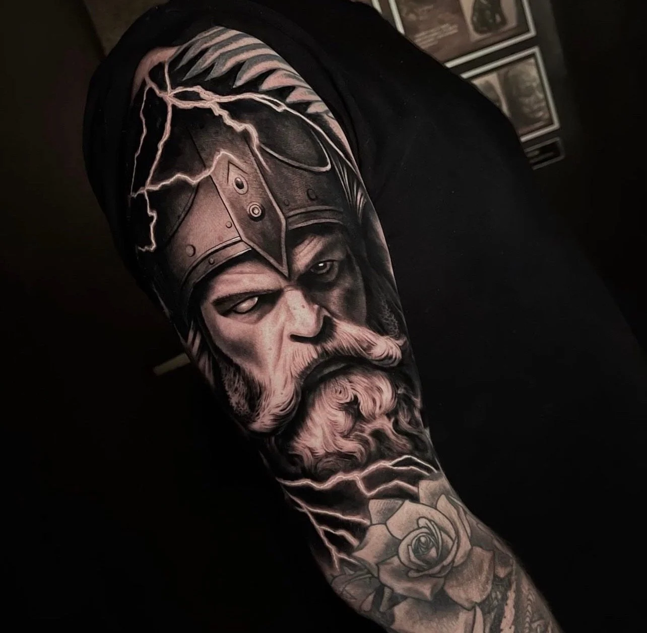 Black and gray tattoo of a fierce Viking warrior with a helmet, beard, and intense expression, decorated with lightning and a rose below.
