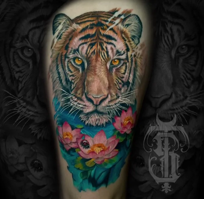Colorful tattoo of a tiger's face above pink lotus flowers on someone's arm.