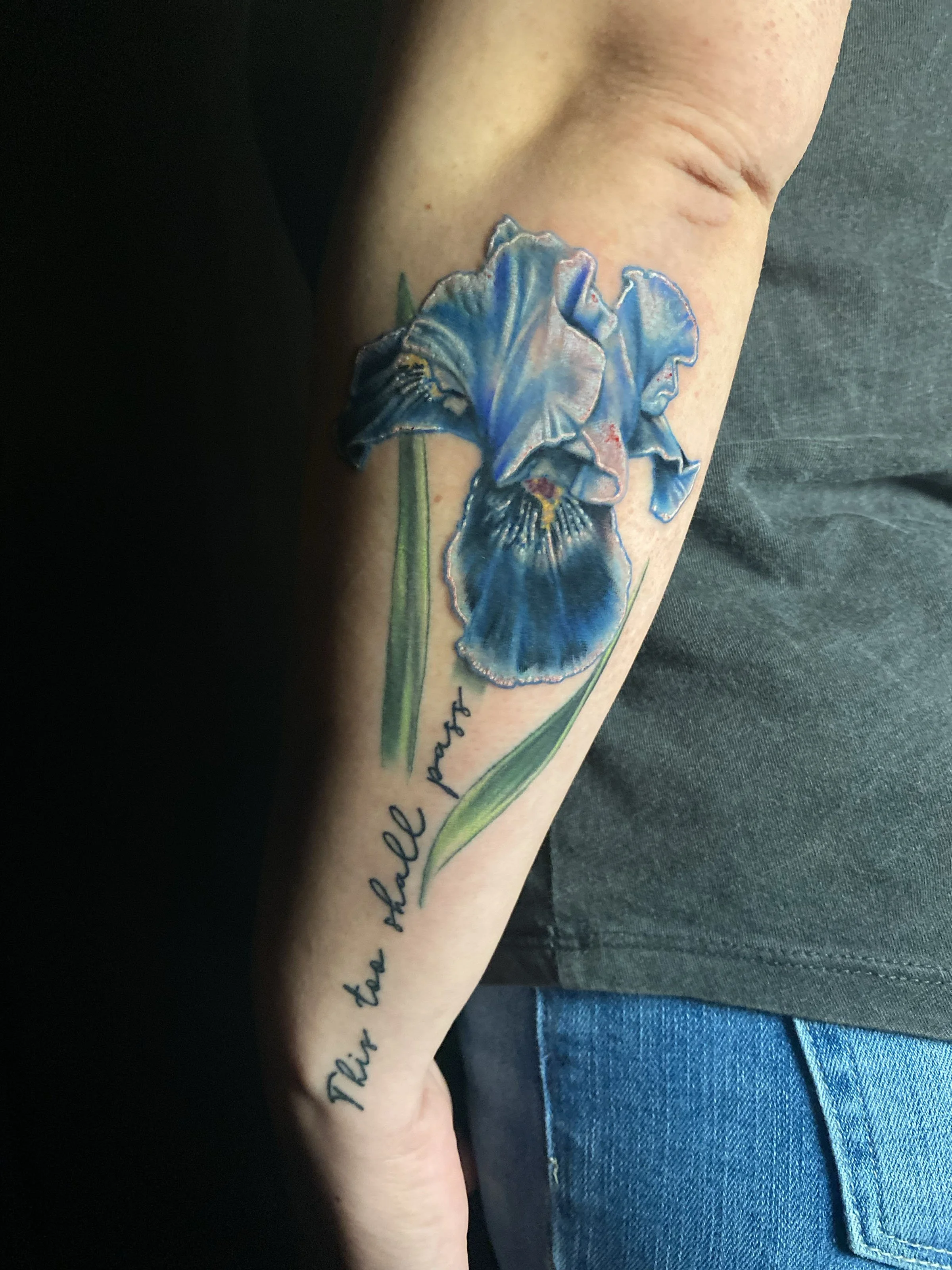Close-up of a person's arm with a colorful iris flower tattoo, accompanied by cursive text.