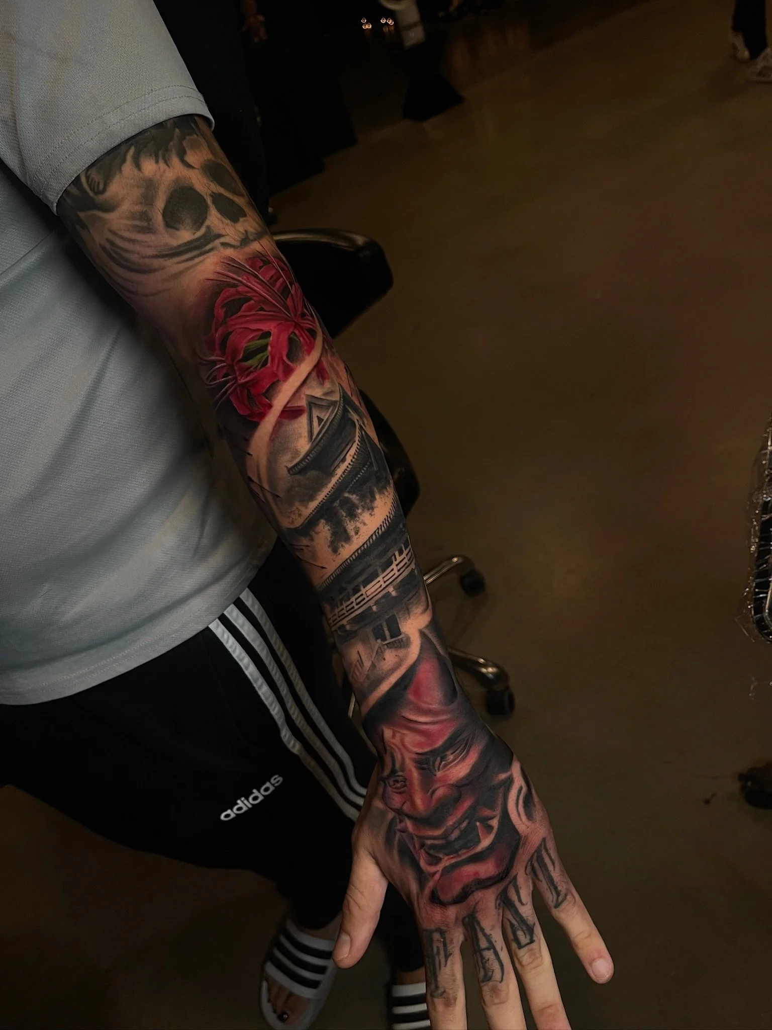 A person with a heavily tattooed arm and hand extended, showing detailed colorful tattoos including a red and green skull, a face with a smile, and various other designs.