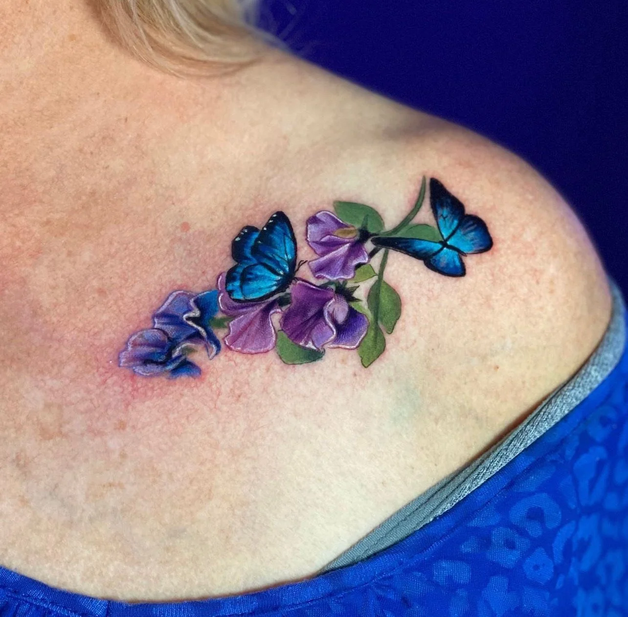 Colorful tattoo of purple flowers with green leaves and blue butterflies on a person's shoulder.
