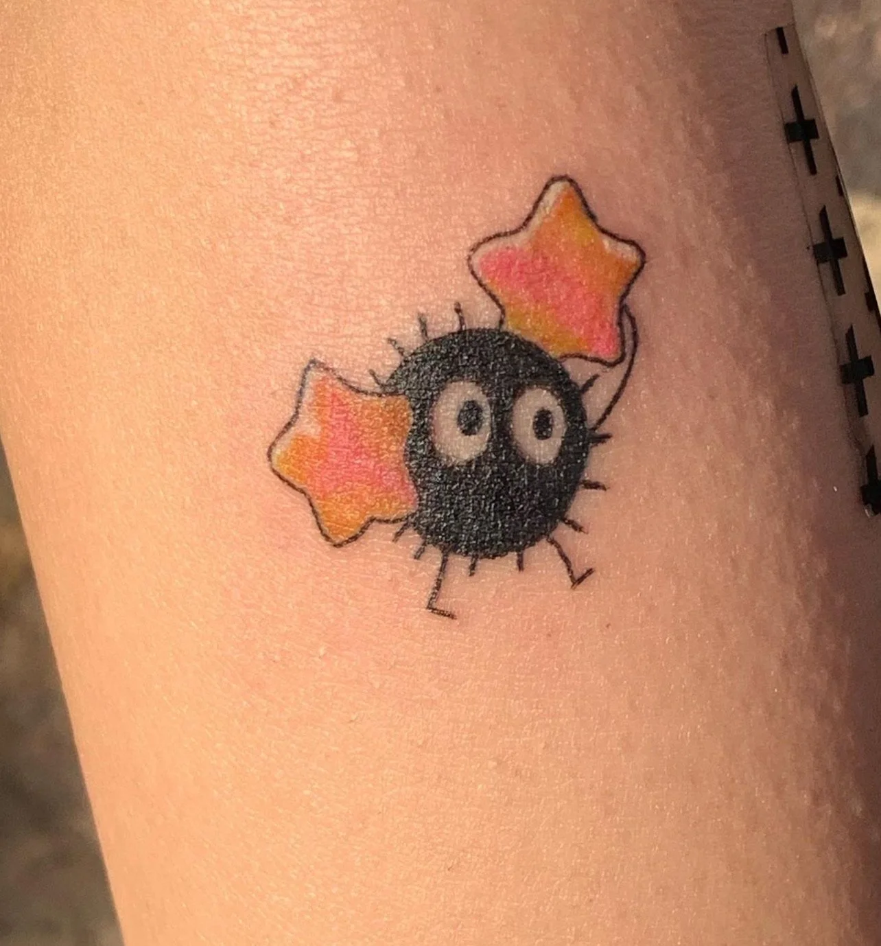 Tattoo of a cartoonish black sphere with white eyes, surrounded by tiny legs and spikes, holding two red and yellow star-shaped objects.