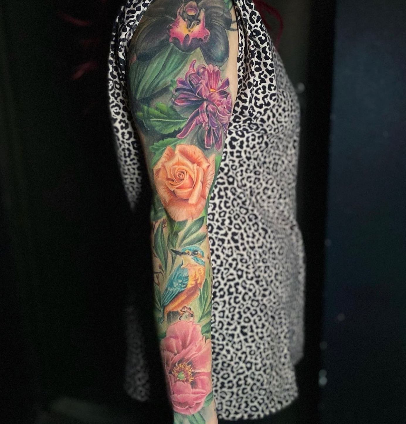 Colorful arm tattoo featuring various flowers including a pink rose, purple flowers, and a pink poppy, along with a small blue and orange bird.