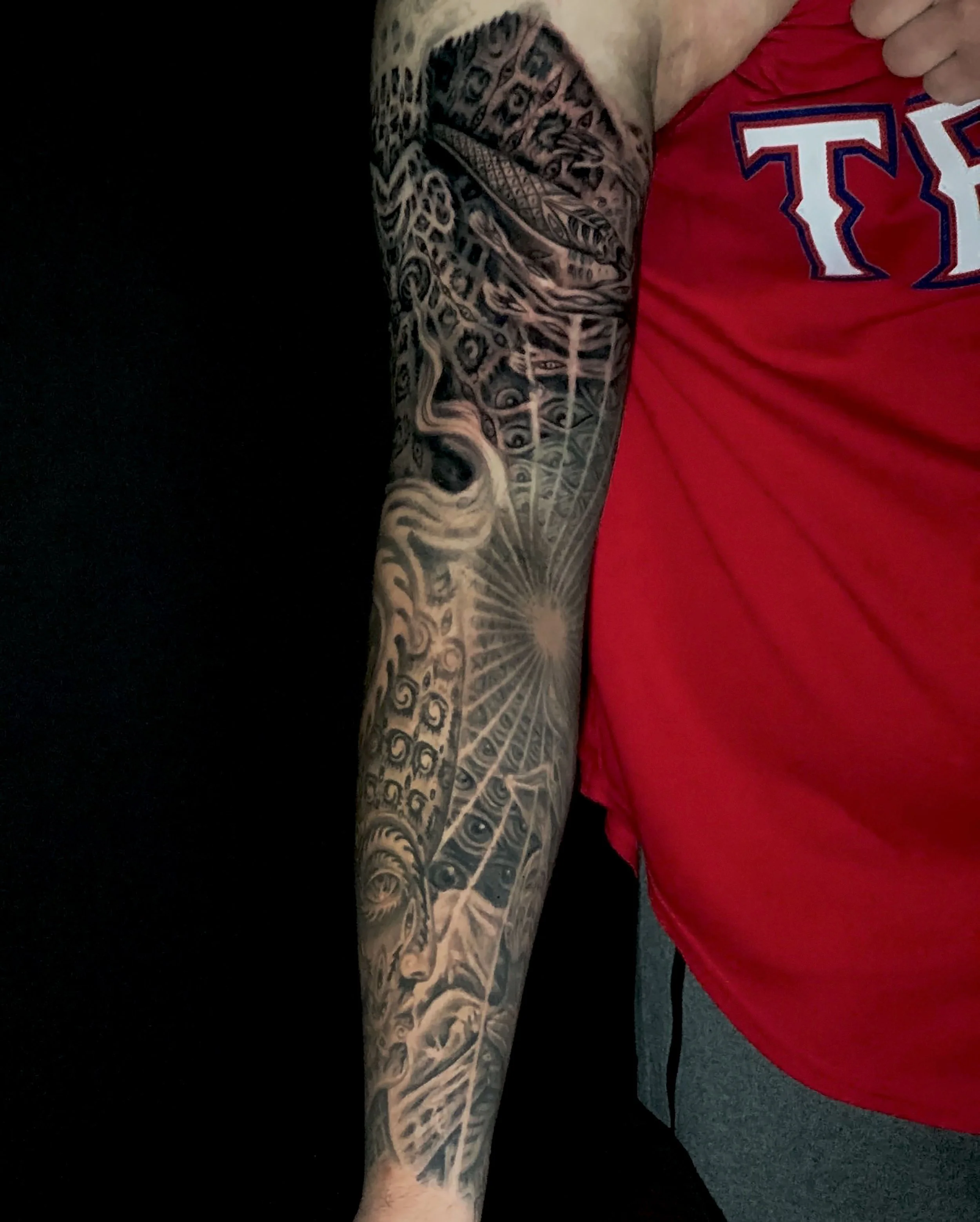 Close-up of a person's arm with a detailed black and gray tattoo, wearing a red shirt with white and blue lettering, seated near black background and gray pants.