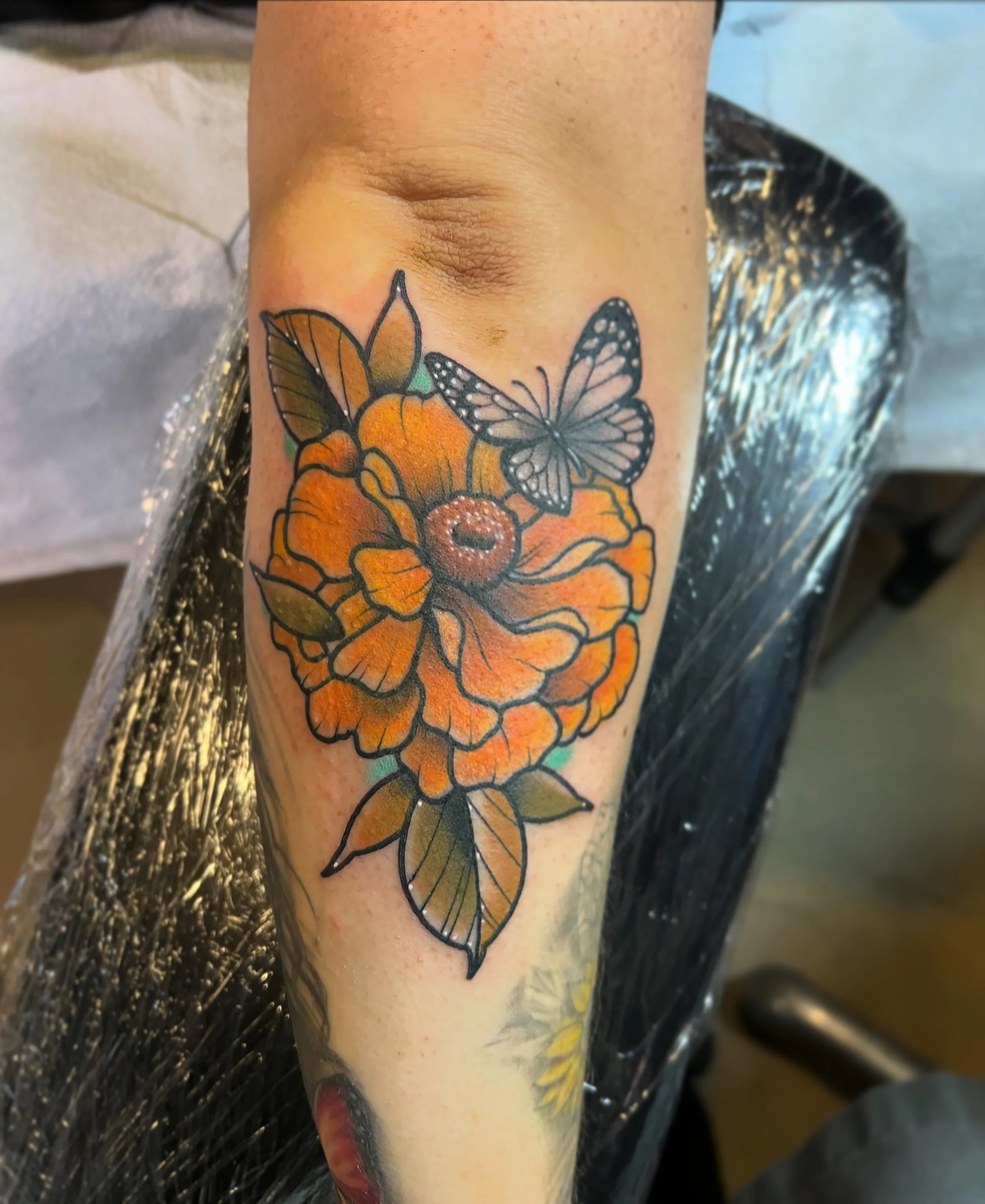 Tattoo of an orange flower with green leaves and a butterfly on top of it on person's forearm.