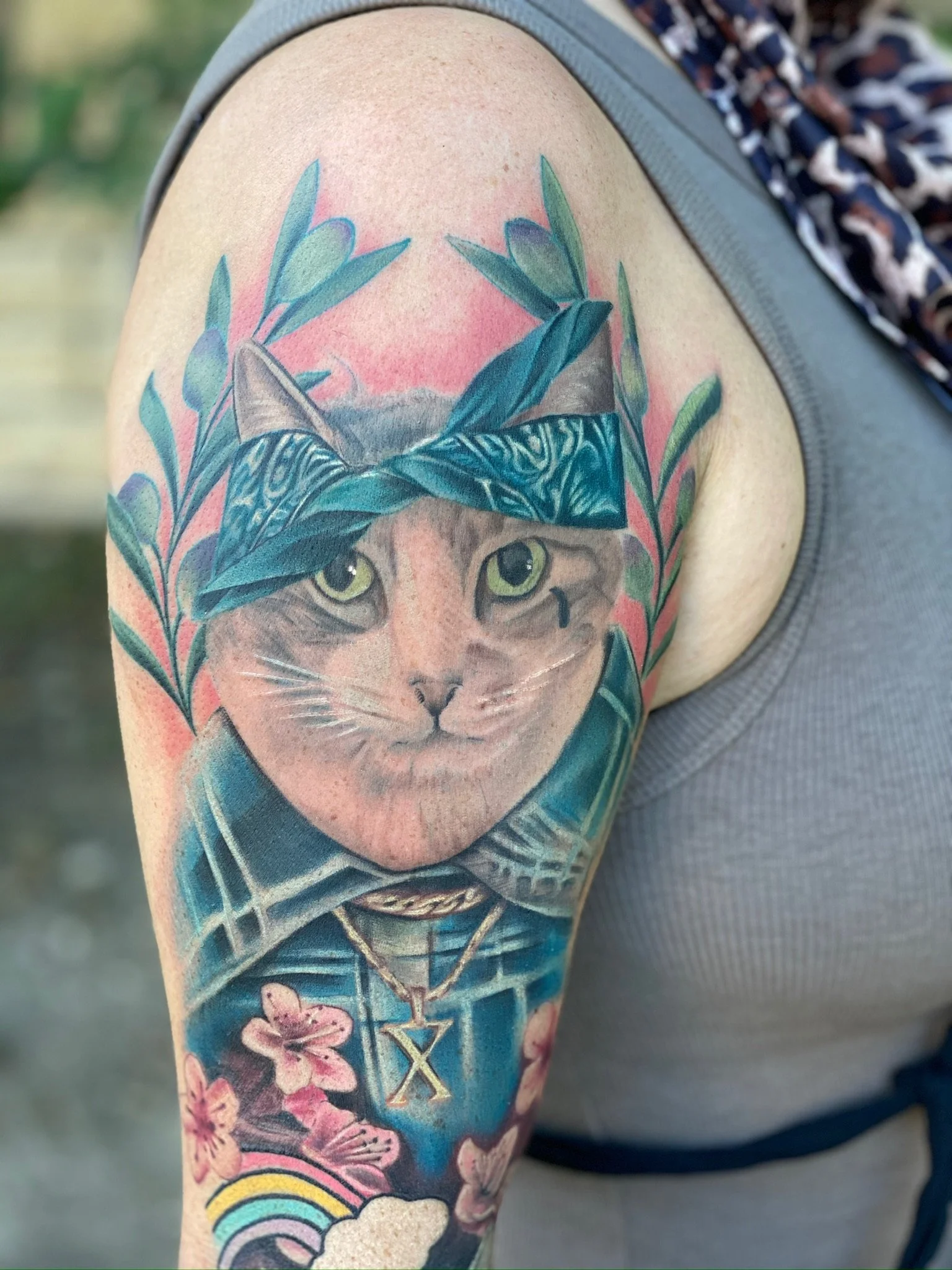 Colorful tattoo of a cat with a bandana on its head, surrounded by greenery, flowers, and a rainbow.