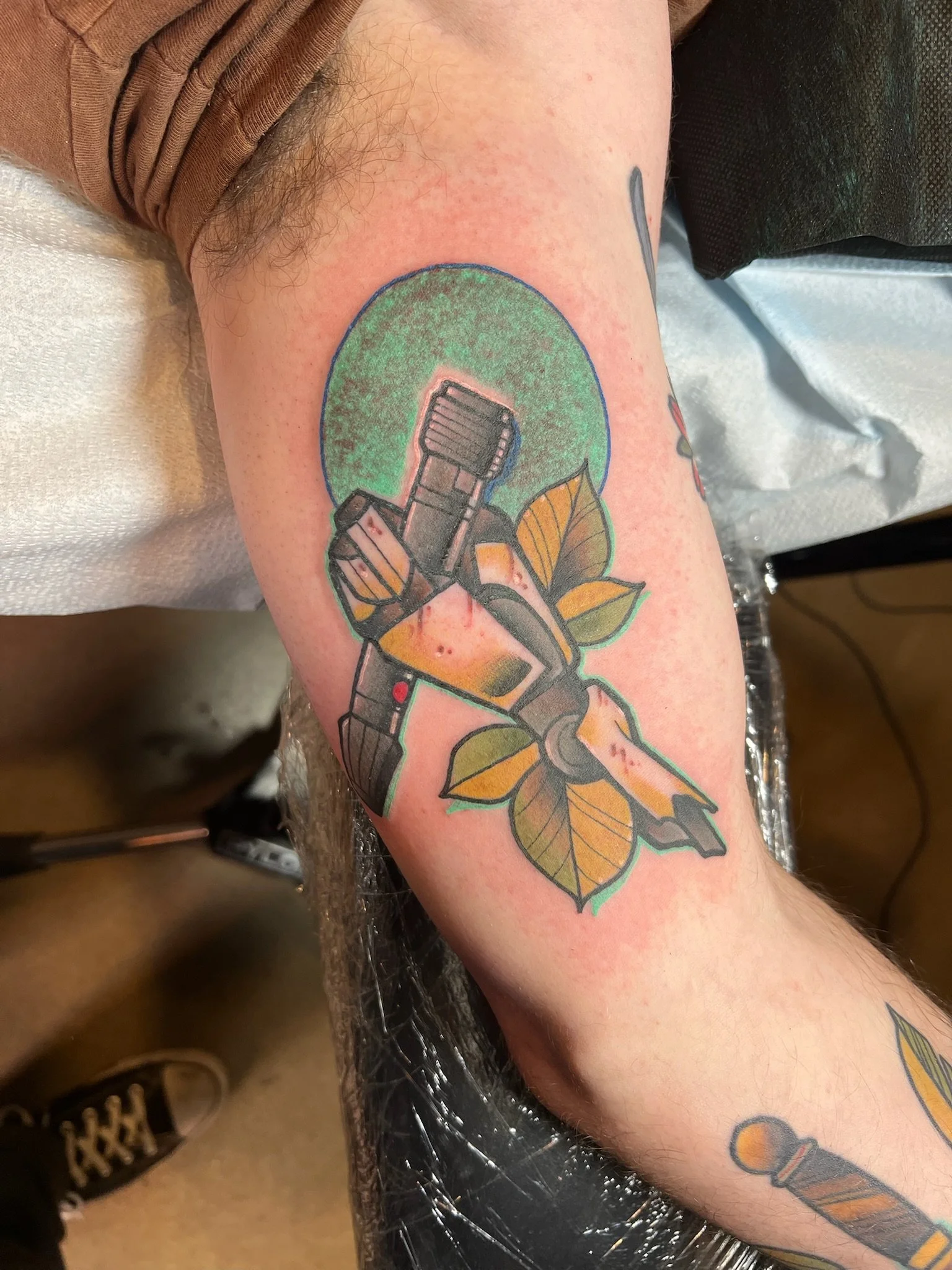 Tattoo of a traditional barber clipper surrounded by leaves, with a green circle in the background.