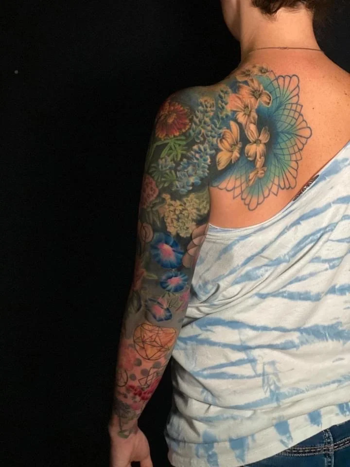 Woman with a colorful floral and geometric tattoo sleeve on her right arm and shoulder, wearing a white and blue tie-dye shirt.