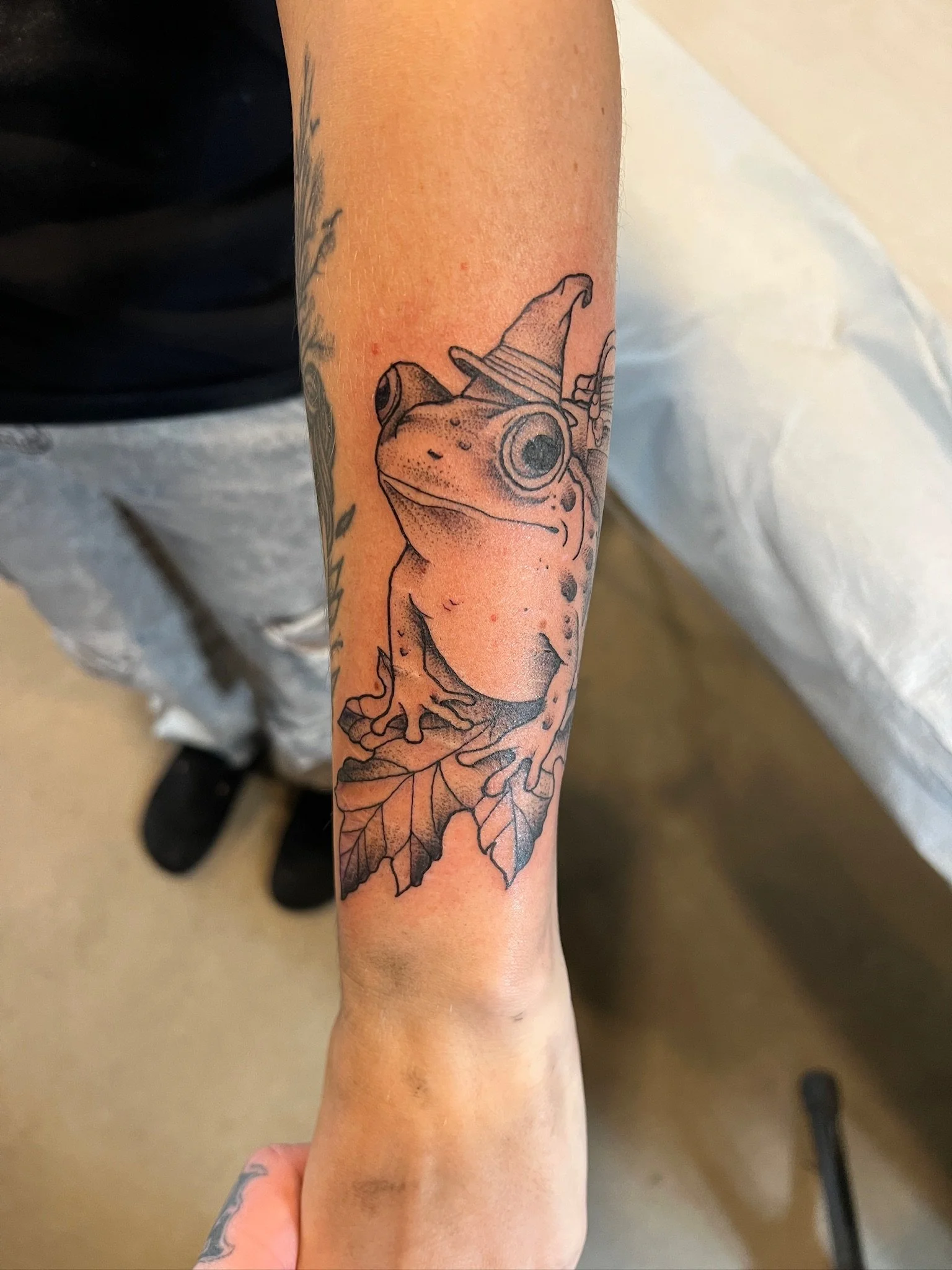 Tattoo of a frog with a witch hat and leaf on the forearm.
