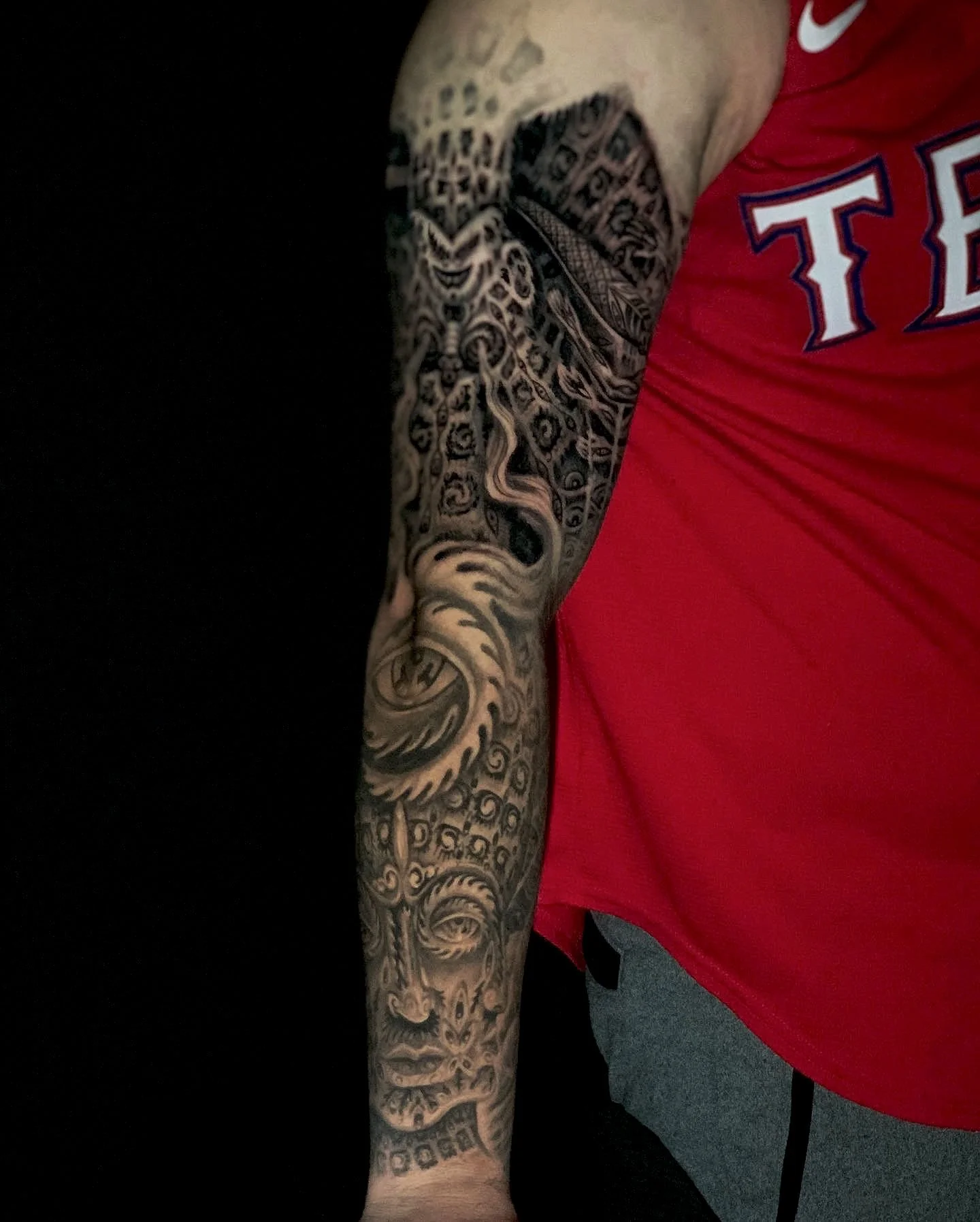 Person with a detailed black tattoo sleeve on their arm, wearing a red shirt with white and blue lettering, sitting against a black background.