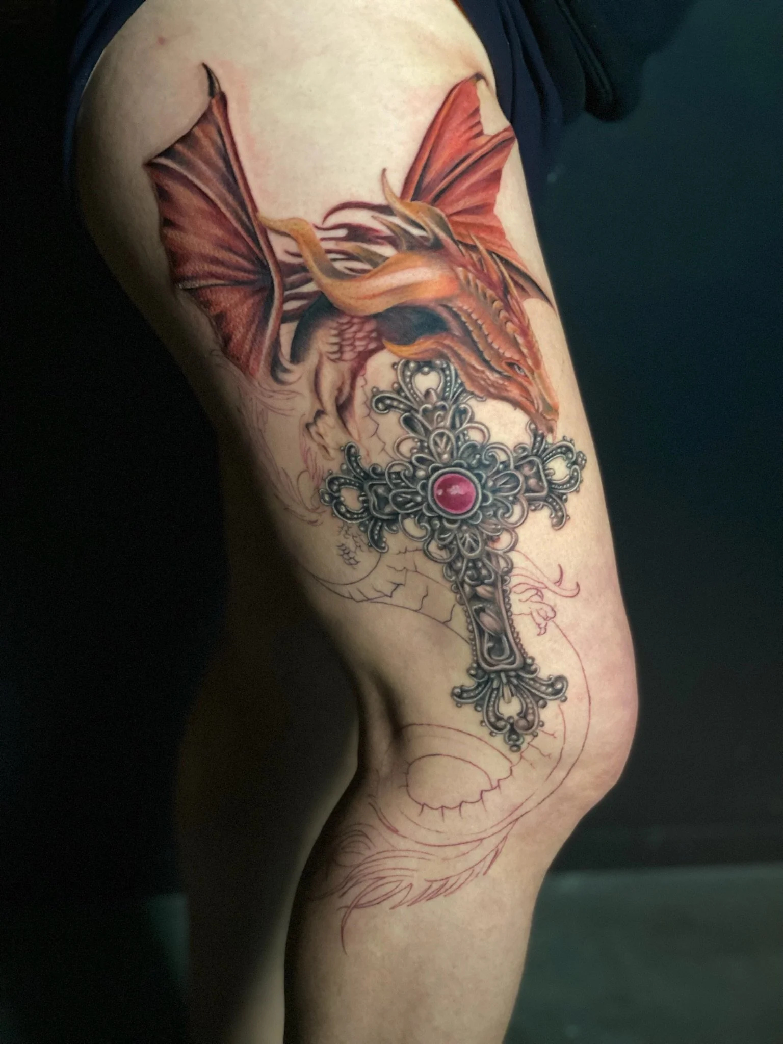 Tattoo of a dragon with red and orange scales and wings, combined with a decorative black and silver cross with a red gemstone in the center, on a person's thigh.