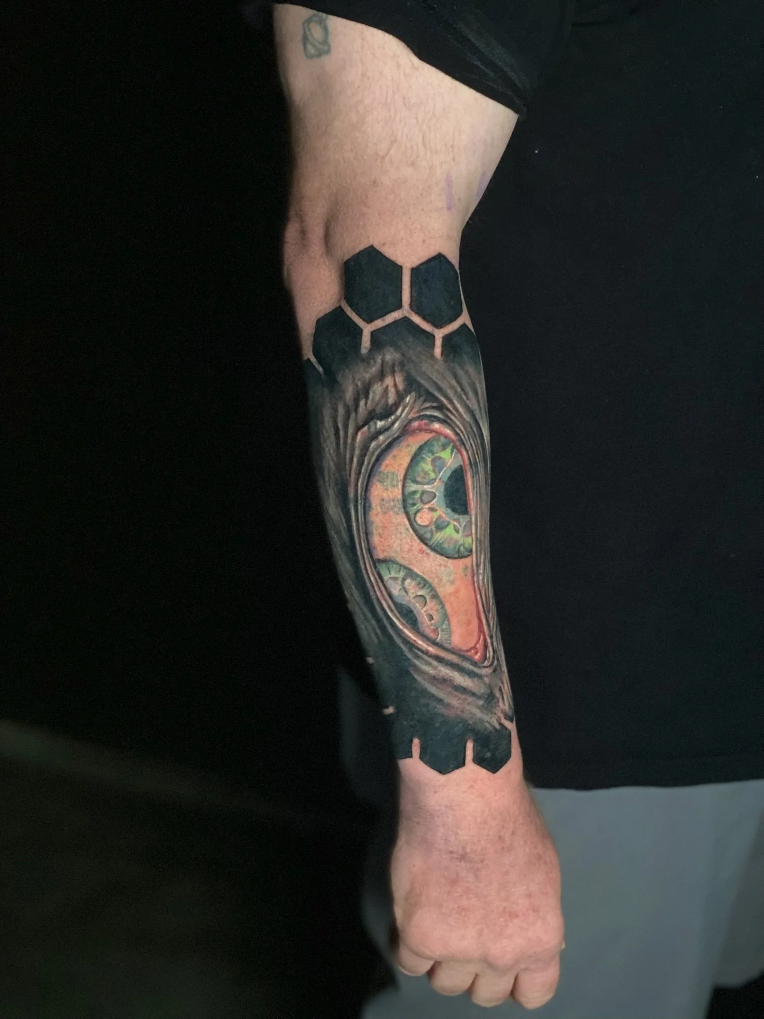 Tattoo of a swirling, spiral eye with vibrant green and orange hues, surrounded by dark shading, with black hexagon pattern extending above the elbow on the arm.