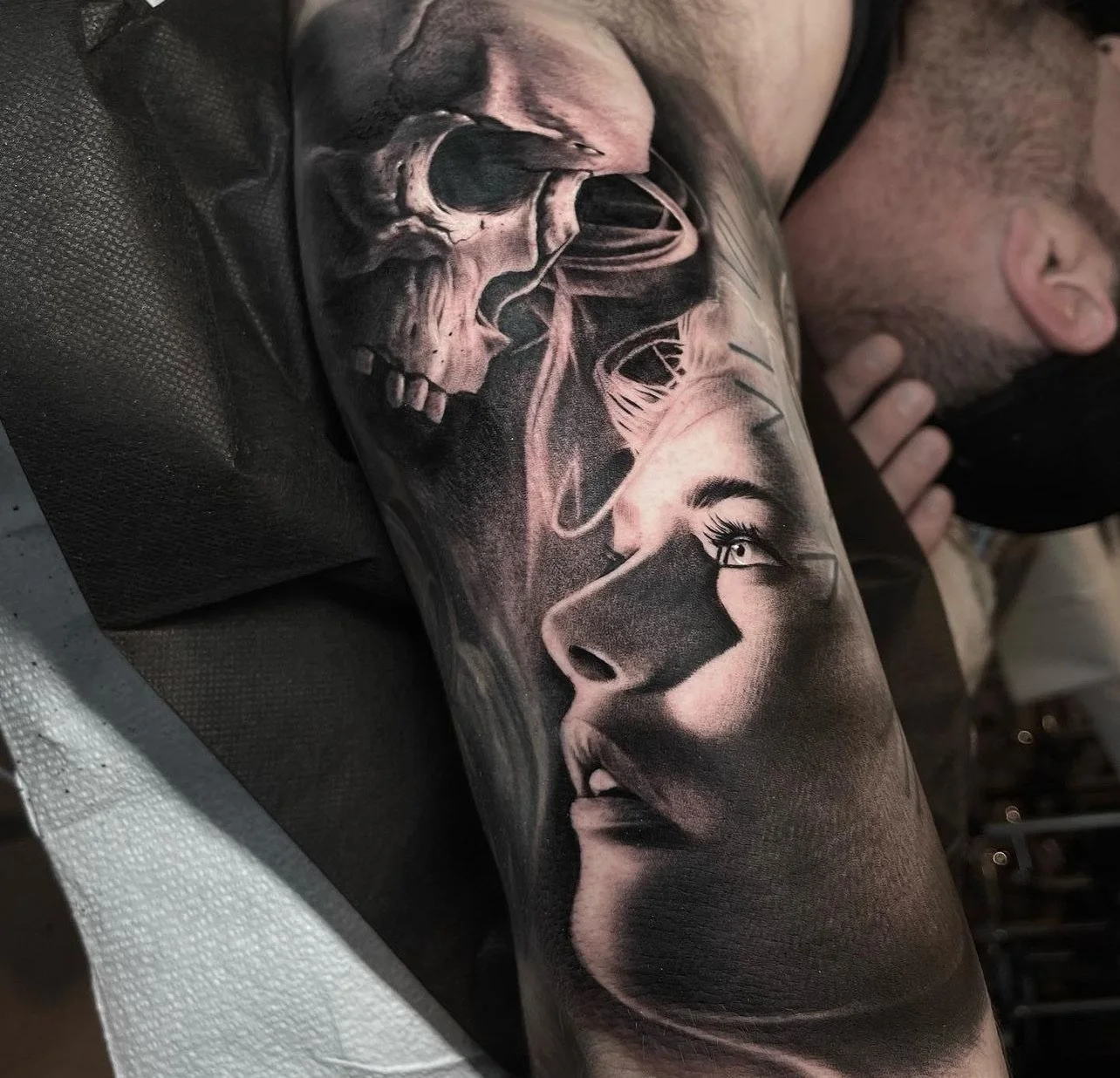 Detailed black and white realistic tattoo of a woman's face with intricate shading on the arm, with a skull and tendrils above her face.