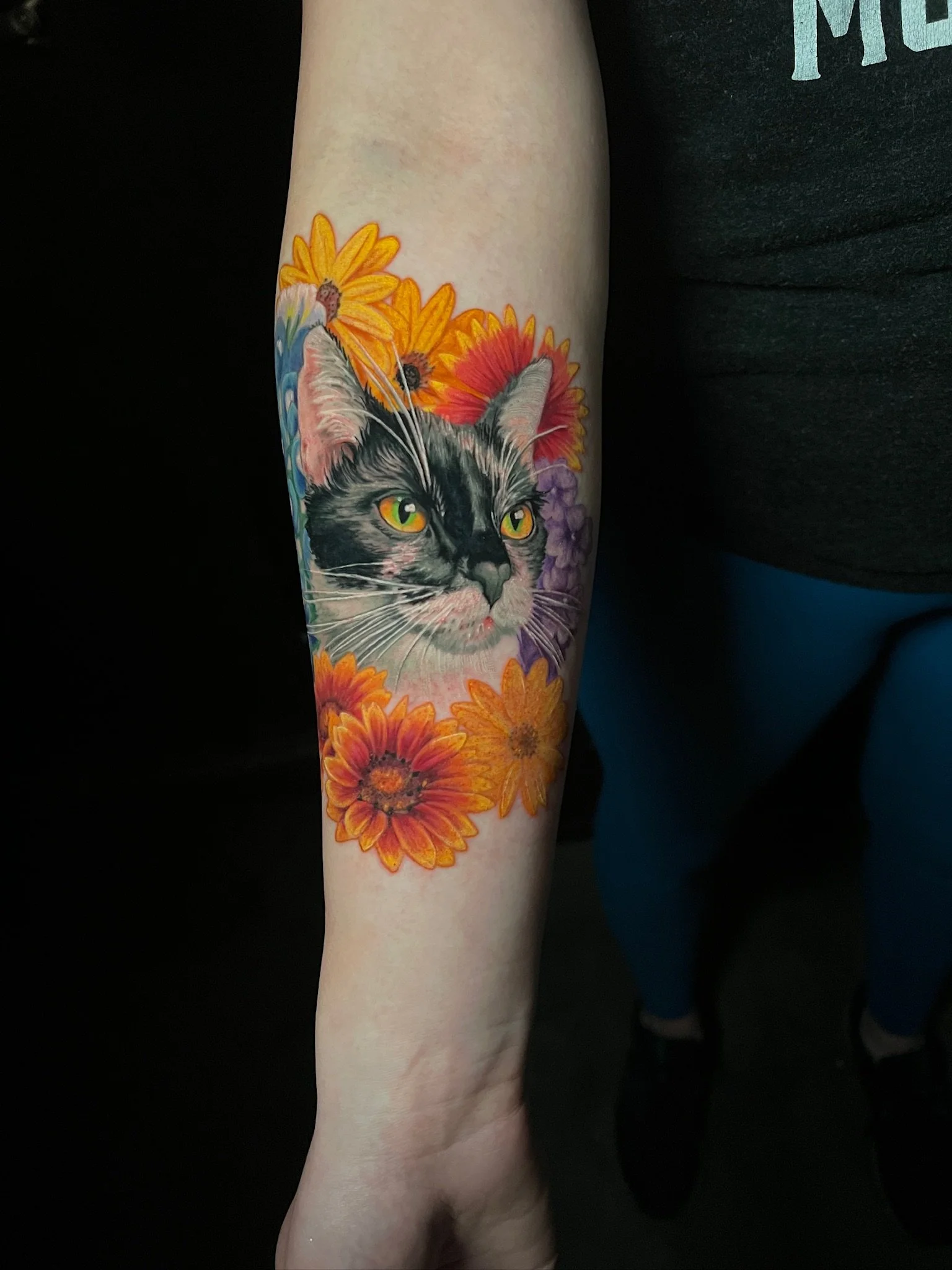 Tattoo of a black and white cat's face surrounded by colorful flowers on a person's forearm.