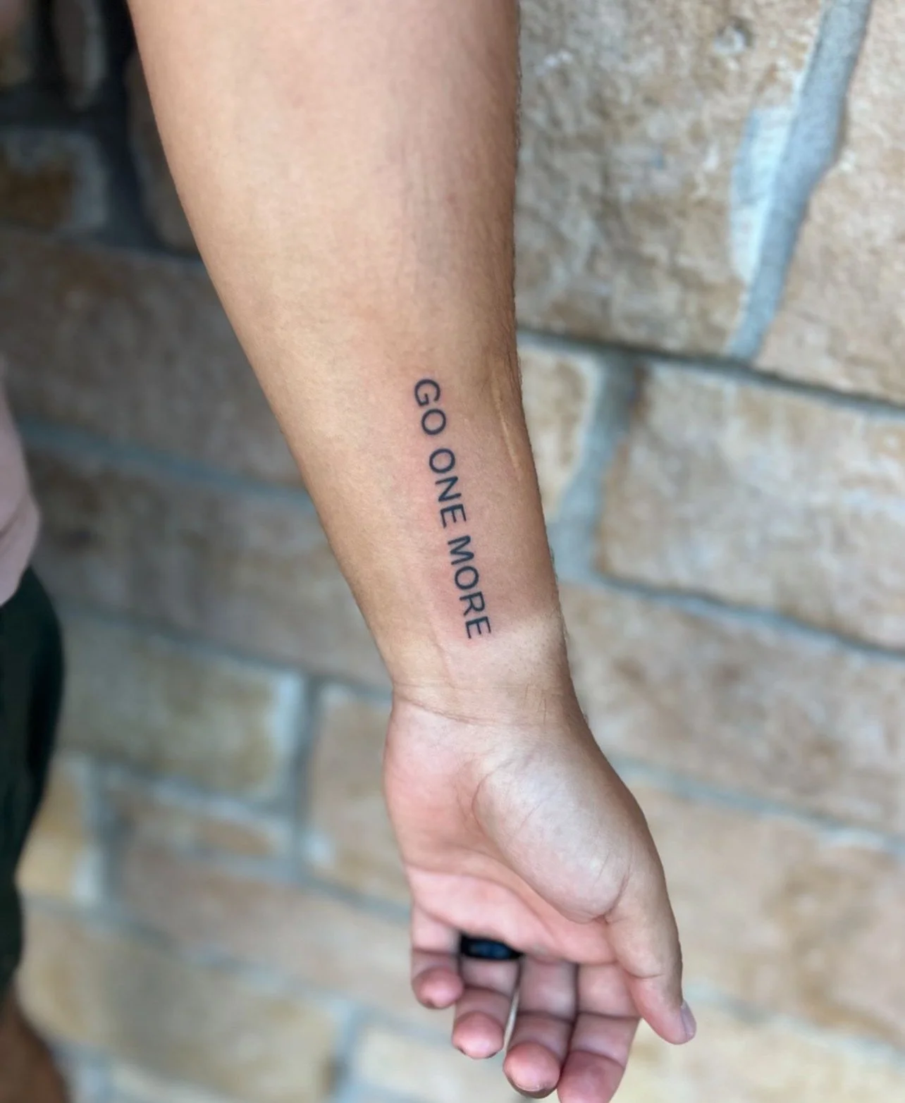 Tattoo on inner forearm with the text 'GO ONE MORE' against a brick wall background.