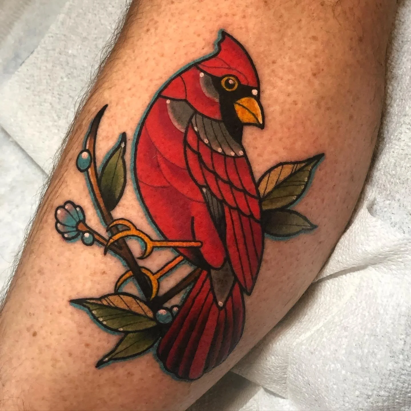 A colorful tattoo of a cardinal bird perched on a branch with green leaves and small berries.