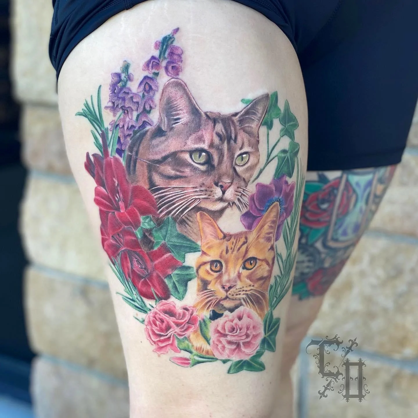 Colorful tattoo of two cats surrounded by flowers on a person's thigh, with one cat above and the other below, featuring detailed fur and expressive eyes.