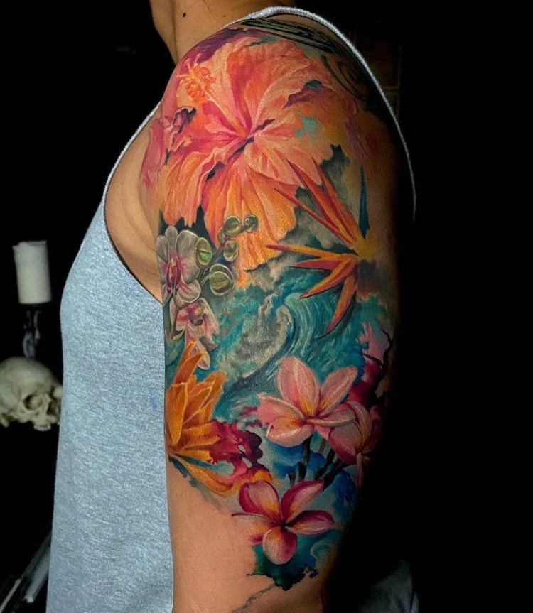 Colorful floral and ocean-themed tattoo on a person's upper arm featuring pink lilies, orange and purple flowers, a wave, and fiery orange leaves.