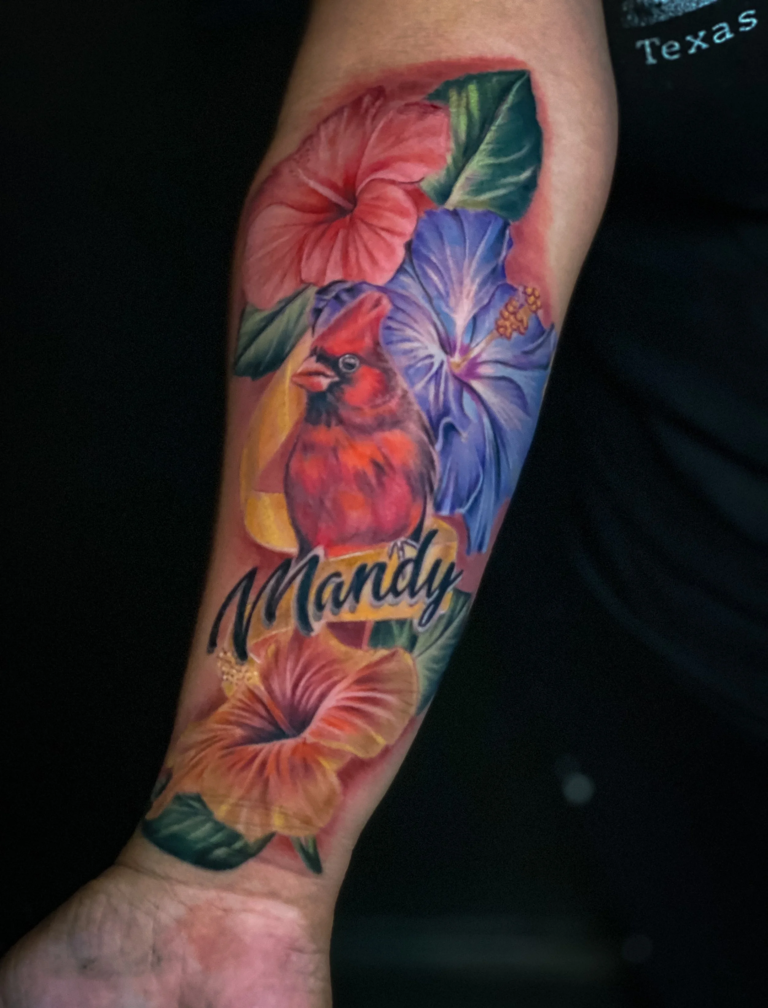 Colorful tattoo on arm featuring a red cardinal bird, hibiscus flowers, and the name 'Mandy' in cursive.