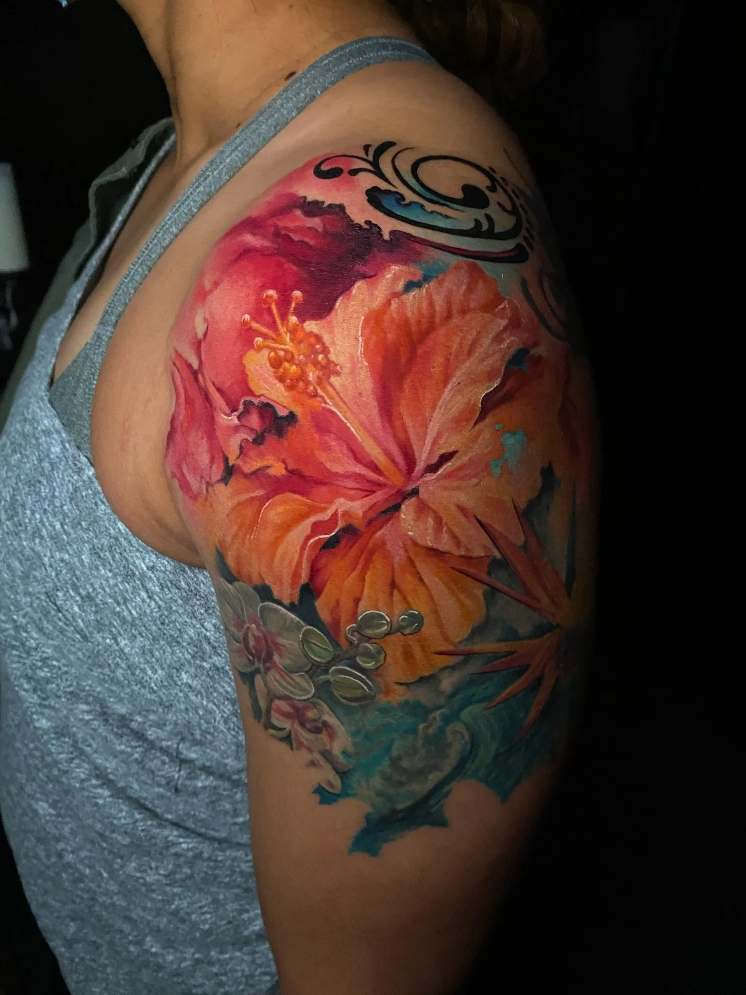 Colorful floral tattoo featuring an orange hibiscus flower, smaller purple and white flowers, and ocean waves on a person's upper arm and shoulder.