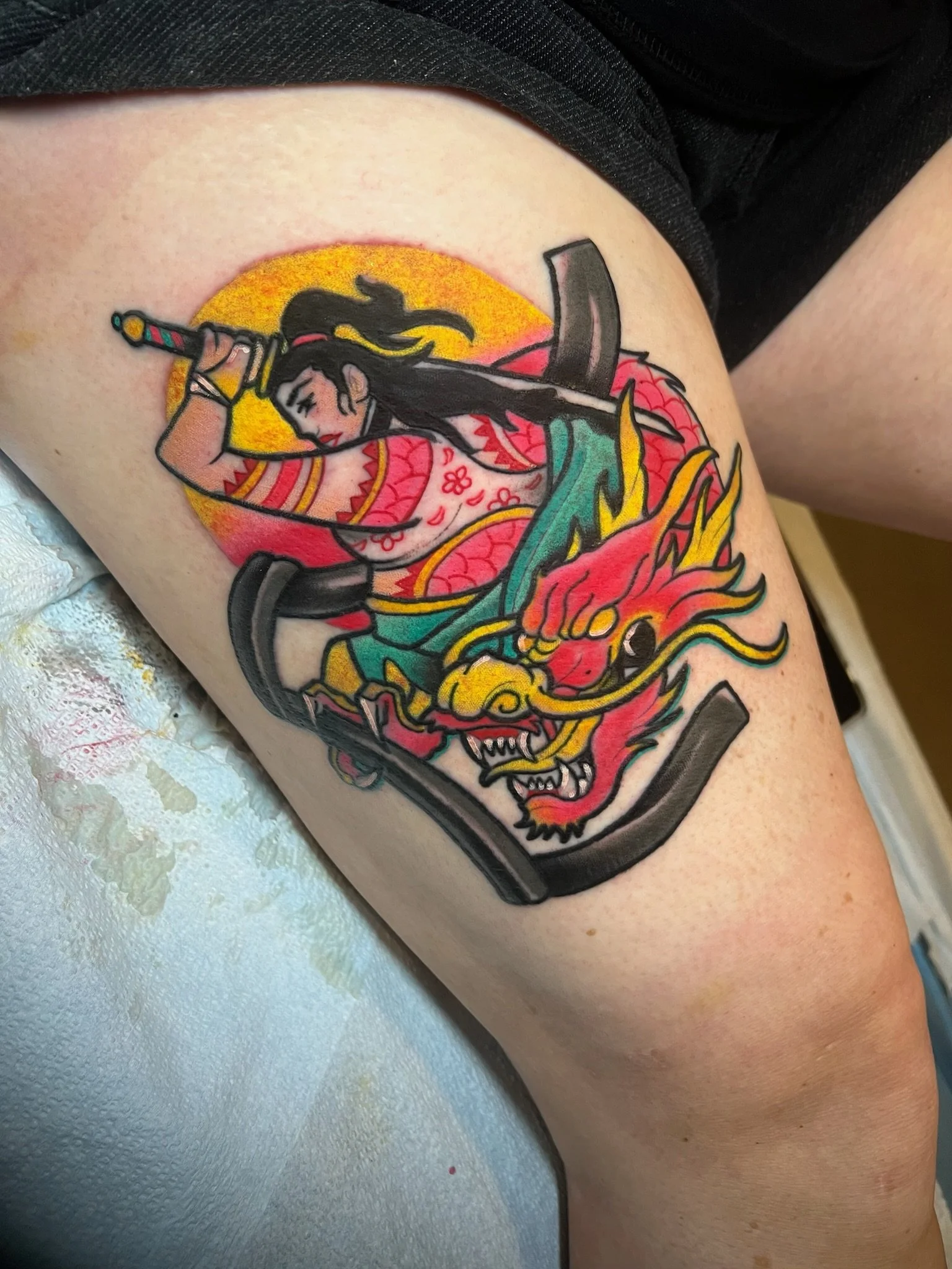 Tattoo of a woman in traditional Japanese clothing wielding a sword, riding a fierce red and yellow dragon, with a yellow sun in the background.