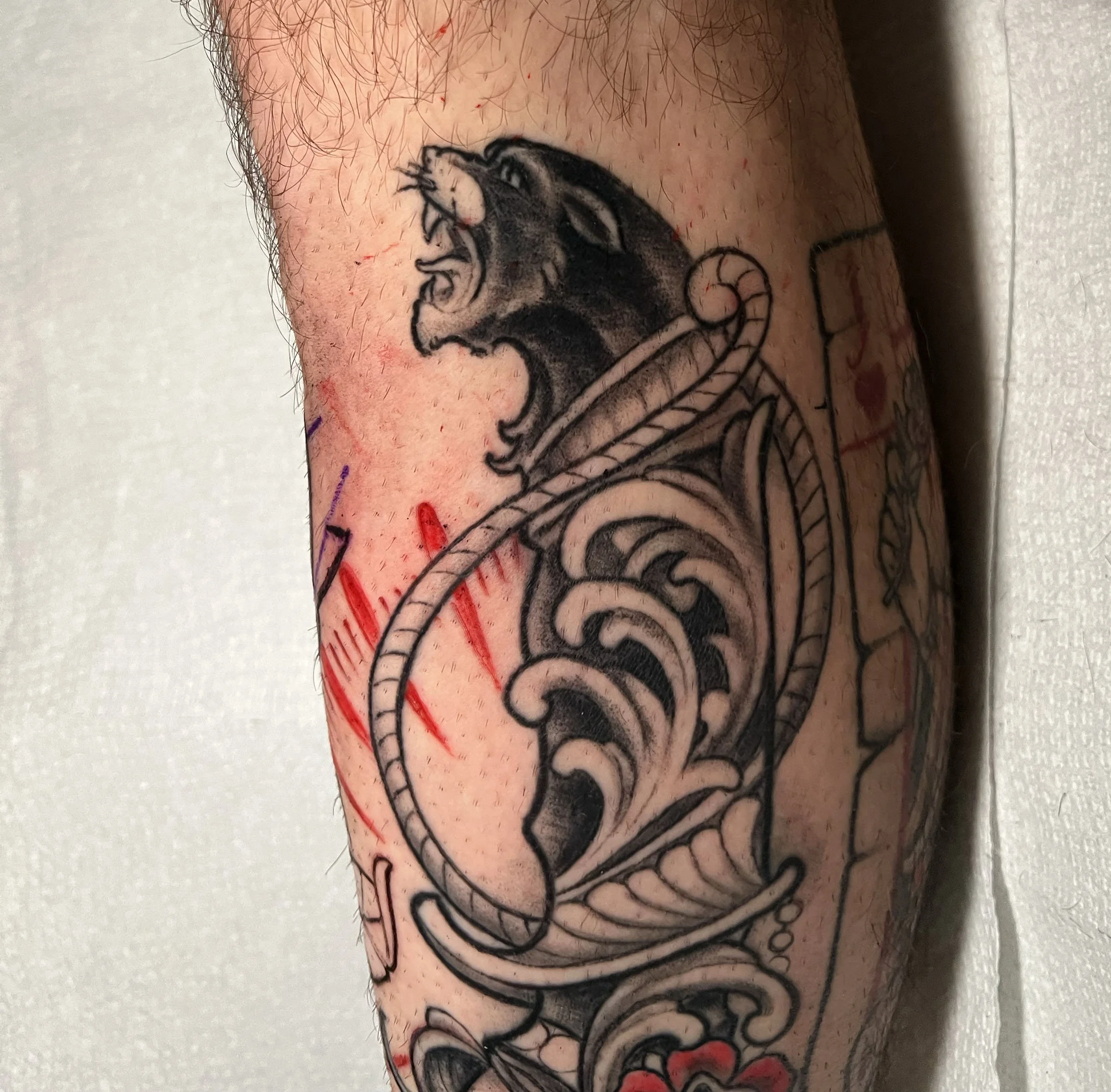 A close-up of a tattoo on a person's arm featuring a black and gray design of a roaring lion's head with intricate details, along with splashes of red ink.