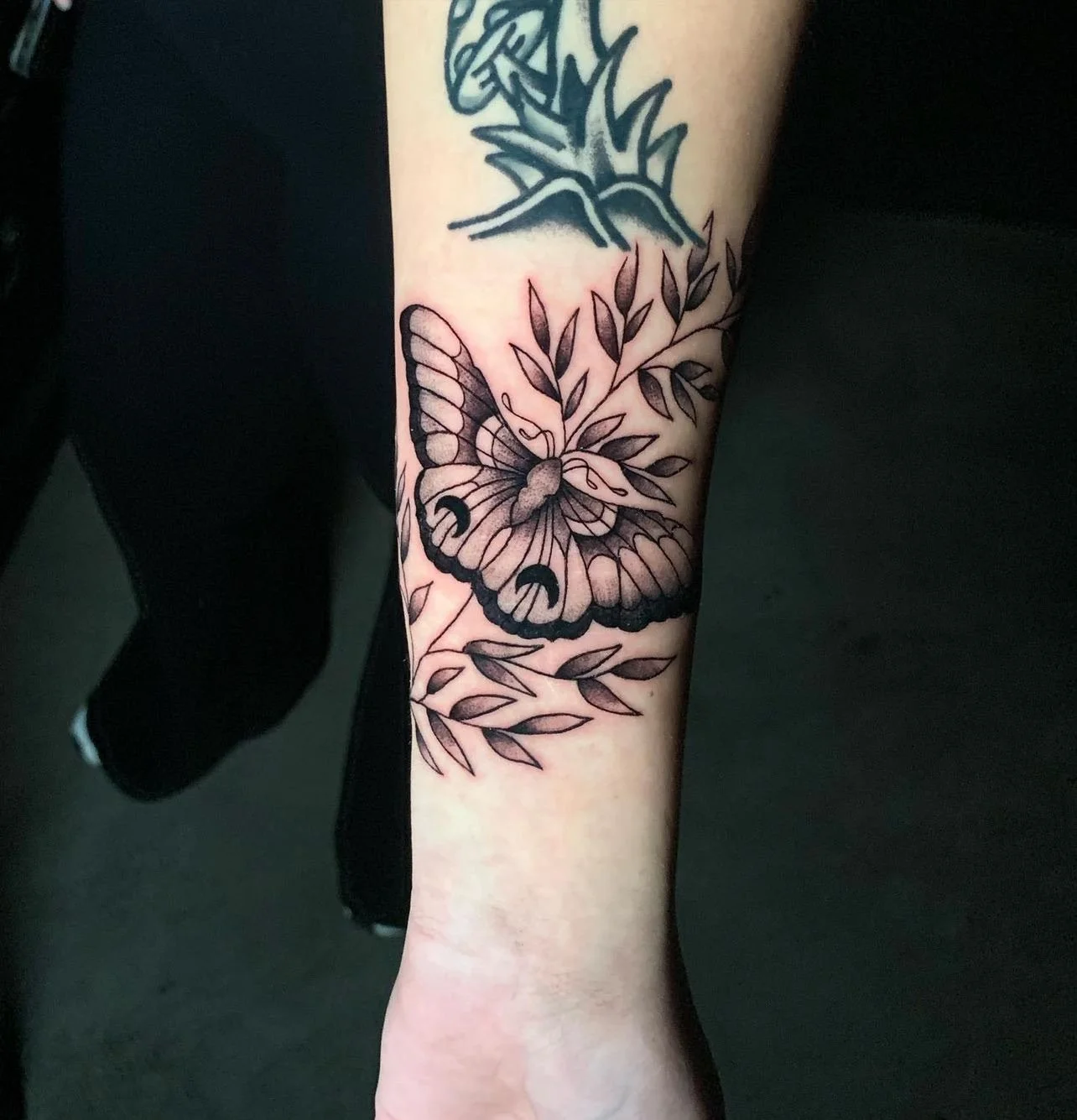 Black and gray butterfly tattoo on person's forearm with leaves and branches.